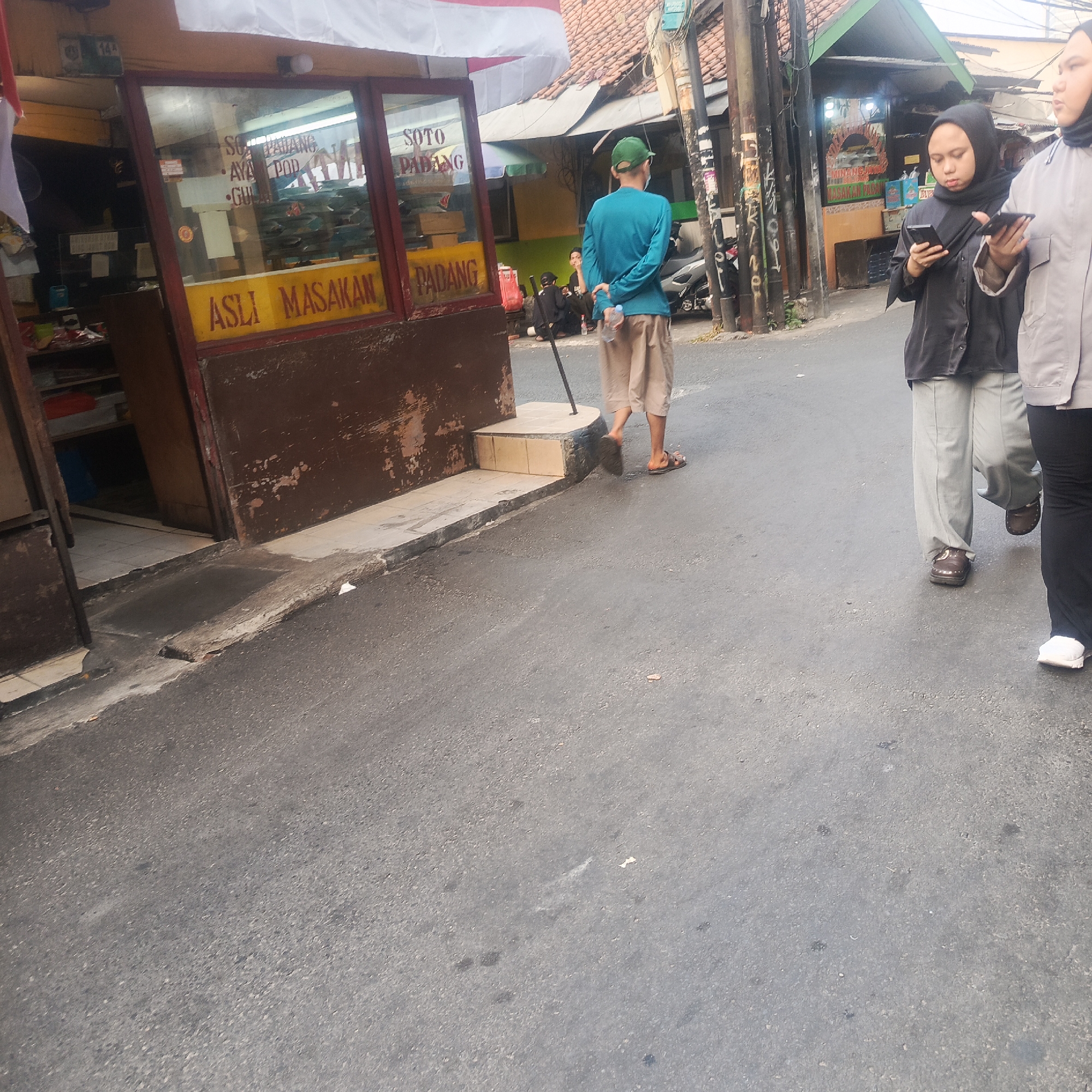 A street-level, medium-distance shot shows a food stall with "SOTO PADANG" and "ASLI MASAKAN PADANG" signs in Indonesian. To the left, a portion of the stall's interior is visible, displaying shelves with various items. In the middle ground, a person wearing a turquoise long-sleeved shirt, tan shorts, and a green cap stands with their back to the camera, holding a plastic water bottle. Further to the right, two women in hijabs walk on the street; the woman on the left is looking at her phone, and the woman on the right is also looking at her phone. Several utility poles stand on the right side of the frame, and a motorcycle is parked behind the food stall. The ground is paved asphalt. The scene appears to be in South Jakarta, Indonesia, based on the style of the food stall and the general environment.