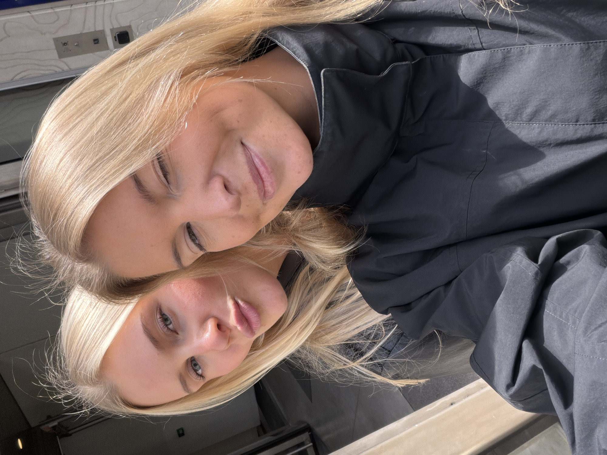 The image is a close-up selfie featuring two women with blonde hair. One woman is positioned directly above the other, their faces aligned. The woman on top has her eyes closed, while the one below has her eyes open. Both have fair skin tones. They are wearing dark jackets. The background includes what appears to be a public transport environment with off-white walls and signage. Given the general aesthetics, it is plausible that the location is a London Underground station, UK.