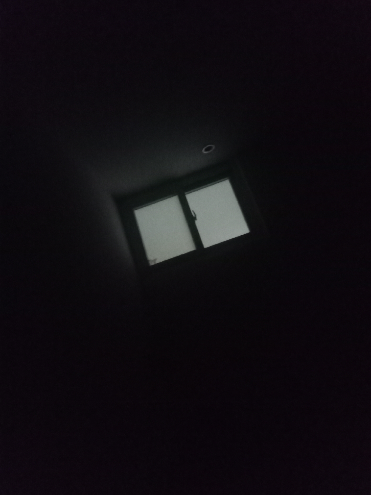 The image shows a dimly lit interior, possibly a room. A window with two panes is visible, allowing faint light to enter. The room is mostly dark, with the window being the primary source of illumination. There is a small, circular light fixture on the ceiling above the window. The exact location is unknown.