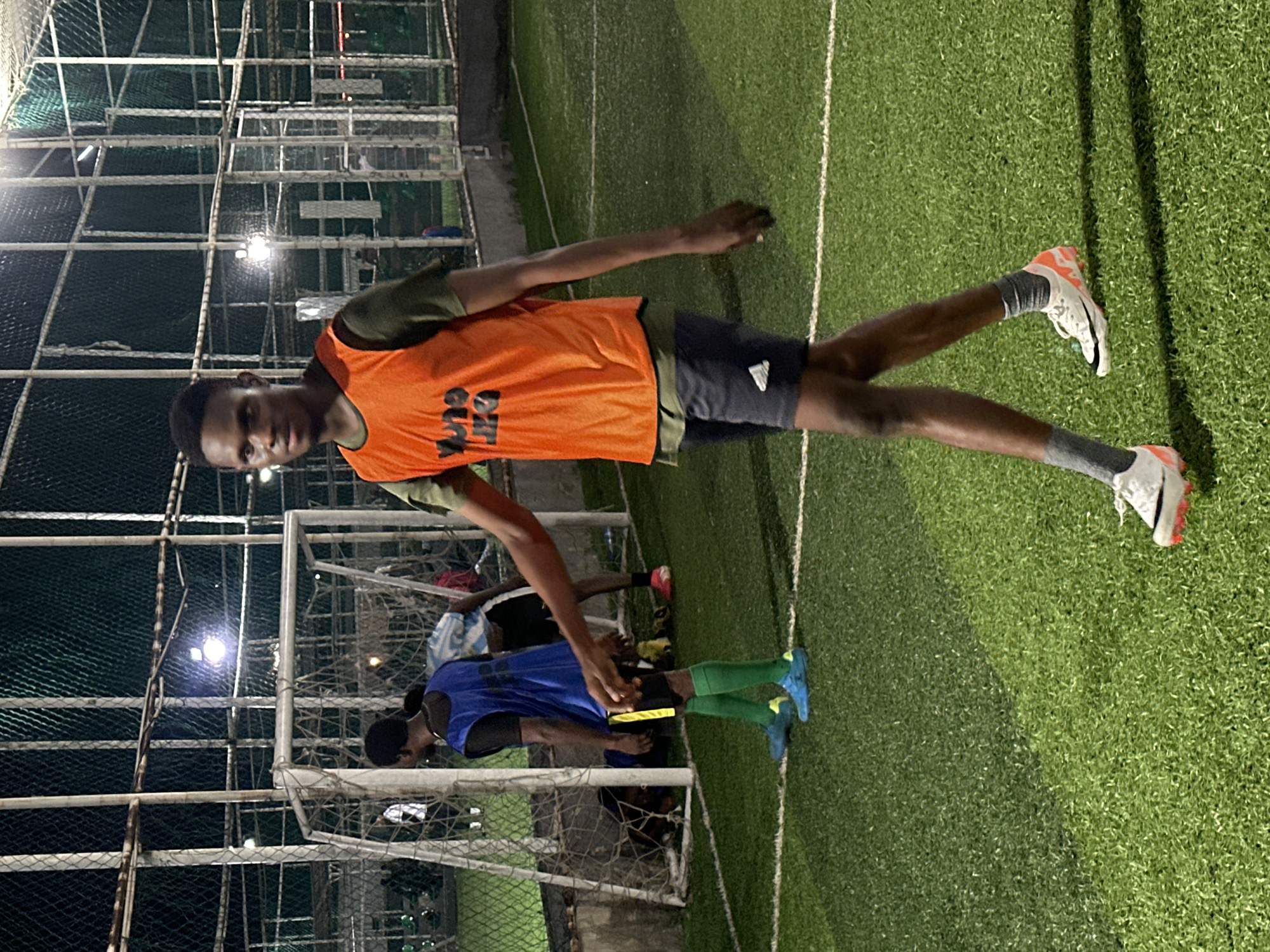 The image depicts an outdoor artificial turf football pitch at night, illuminated by stadium lights, likely in Abuja, Nigeria. A young Black male, wearing an orange bib over a dark green t-shirt and dark shorts, walks towards the viewer. He has short, dark hair and is looking slightly to his left. His feet are clad in white and orange soccer cleats and grey socks. In the background, a soccer goal and net are visible, as are other individuals in athletic wear, some in blue and white jerseys. The overall scene suggests a training session or informal game.
