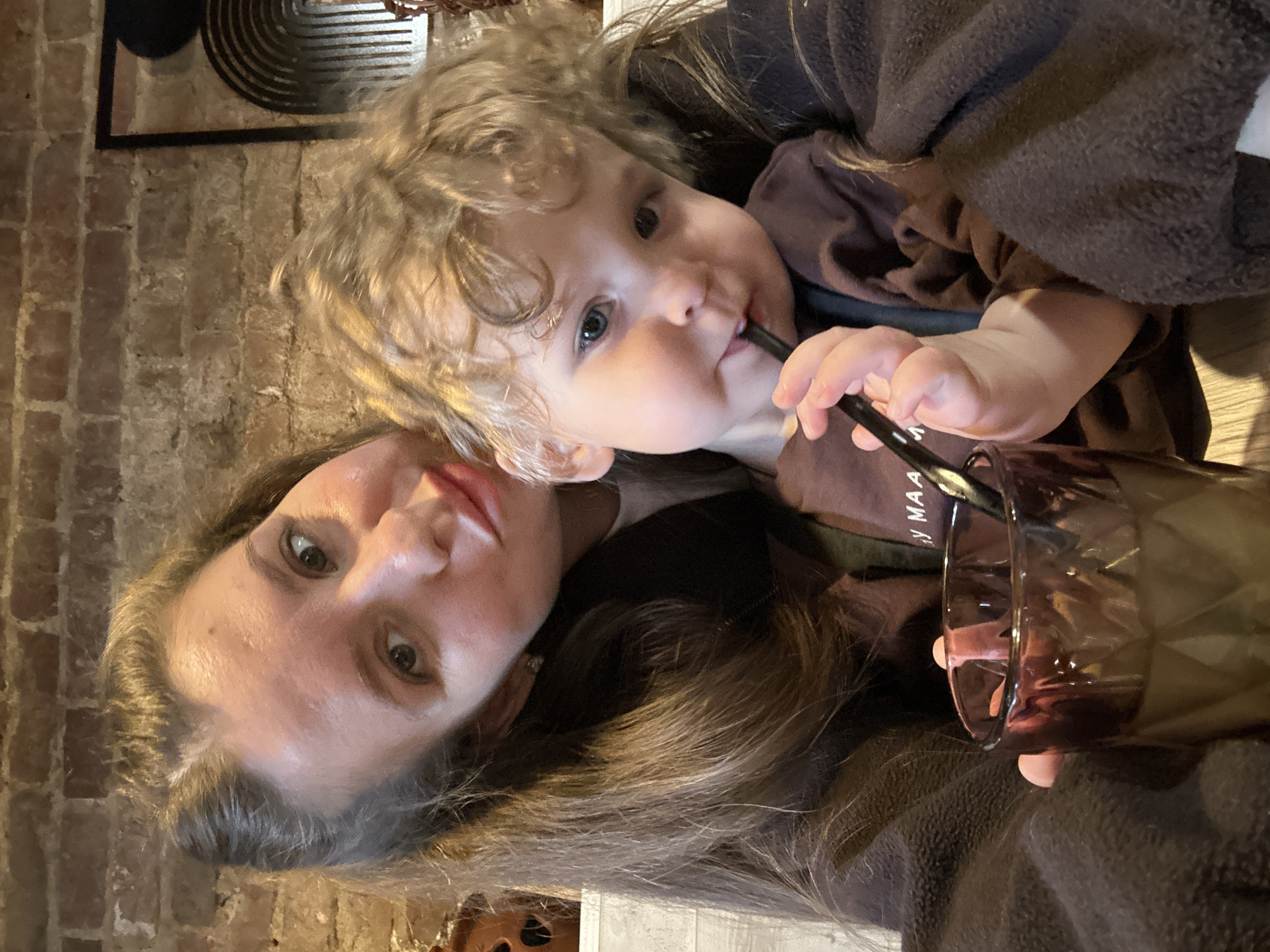 A woman with long brown hair and a child with curly blond hair are in the image. The child is drinking from a glass with a black straw. The background includes a brick wall and a decorative metal piece. The setting appears to be indoors. Klin, Russia is not discernable from this image.