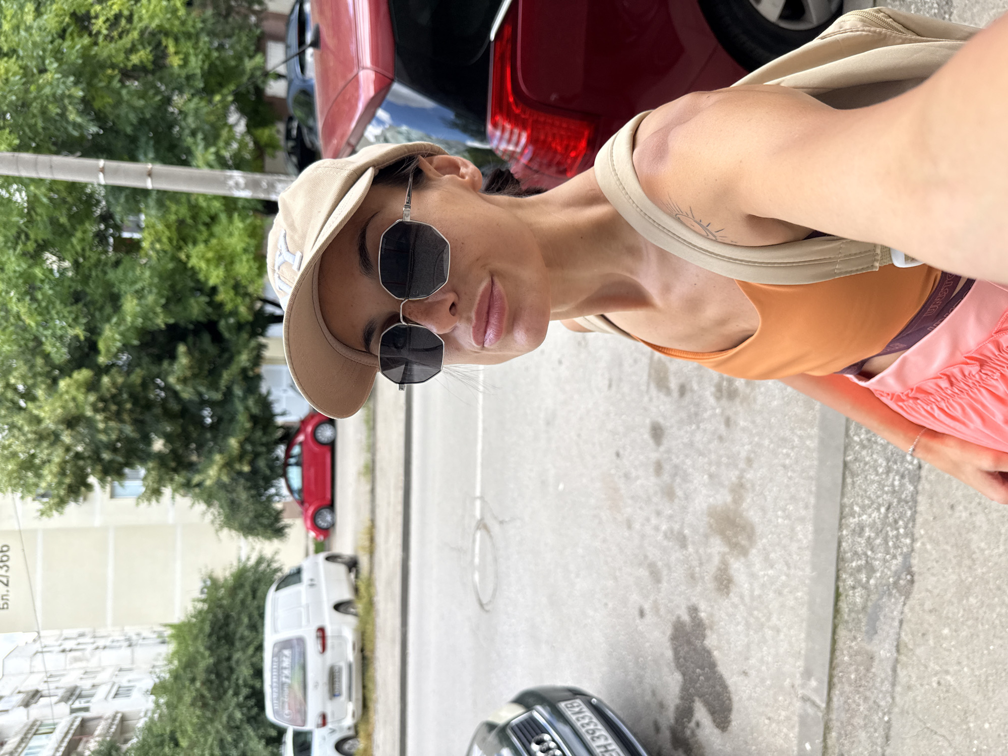 A woman, wearing an orange tank top, pink shorts, a beige baseball cap, and sunglasses, stands on a sidewalk next to a street. Behind her, a red car is parked. In the background, there are trees, buildings, and other cars, suggesting an urban environment. The image appears to have been taken in Pleven, Bulgaria, based on the building number visible in the background which reads "бл. 2/366".