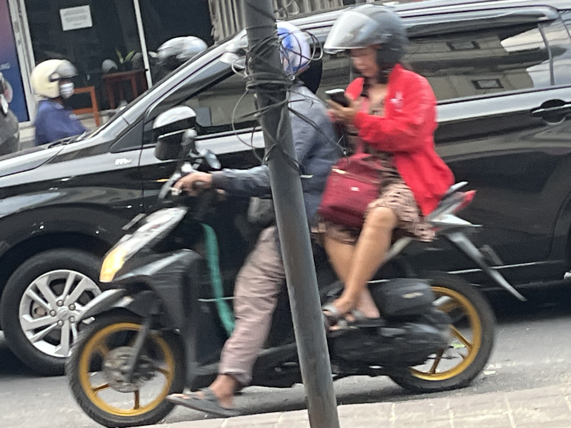 In North Kuta, Indonesia, two people are riding a scooter on a paved road. The person in front, the driver, is wearing a grey helmet and a blue jacket, and appears to be focused on the road ahead. The person in the back is wearing a red jacket and a black helmet, and is looking at their smartphone. They are carrying a red bag on their lap. A black car is visible to the left of the scooter, and parts of a building and a street light are also in the frame. The scooter has a black body and gold wheels. The overall scene suggests a daytime commute in an urban environment.