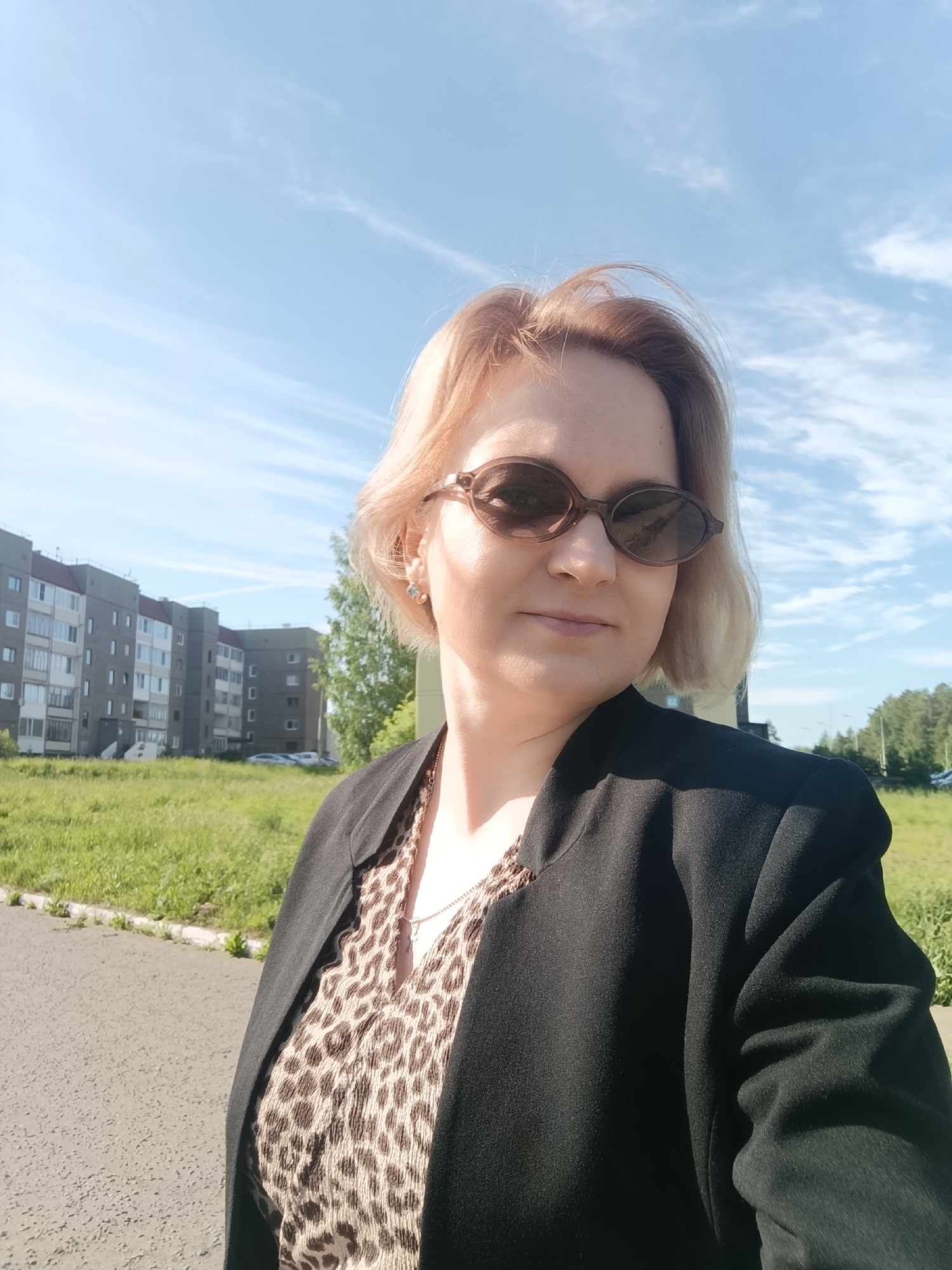 A woman with light blonde hair and sunglasses is the central subject of this outdoor half-body portrait. She is wearing a black blazer over a leopard print top, with a silver chain visible around her neck. Her expression is serene, with a slight, subtle smile.
The setting is an open area, likely a residential neighborhood, on a bright, sunny day. In the background, several multi-story apartment buildings with balconies are visible, fronted by a large expanse of vibrant green grass. A paved path or road runs across the foreground and mid-ground. Trees are also present in the background, beyond the buildings. The sky is a clear, bright blue with scattered wispy white clouds. The strong, natural sunlight suggests it is daytime, likely mid-morning or afternoon. The architecture of the buildings is consistent with the provided location context of Марковский, Russia. No visible text is present in the image.