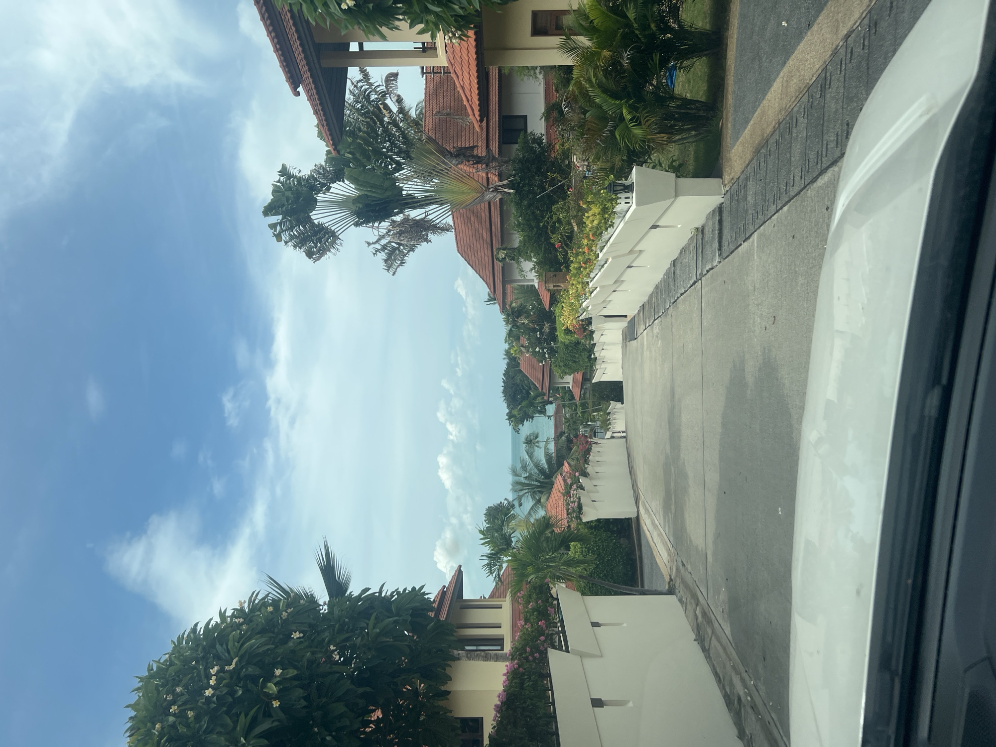 The image shows a downward sloping street in a residential area on Ko Samui, Thailand. The street is paved with concrete and has a white railing on either side. Lush tropical vegetation, including palm trees and flowering bushes, lines the street. In the background, several houses with terracotta tile roofs are visible, nestled among the greenery. Beyond the houses, the ocean can be seen under a bright blue sky with scattered clouds. The perspective suggests the photo was taken from inside a vehicle, as the hood of a white car is visible in the foreground.