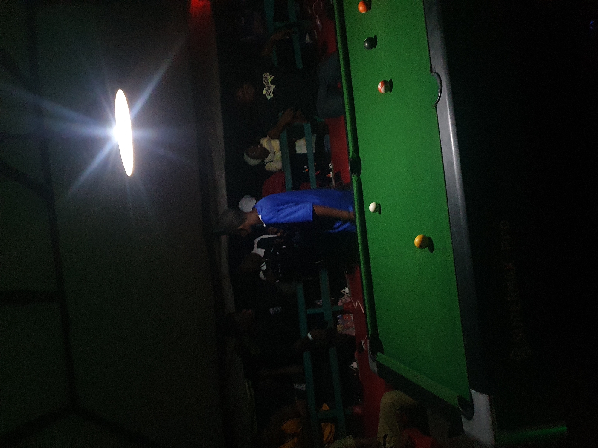 The image depicts a dimly lit indoor scene, possibly a bar or recreational center, in Lokoja, Nigeria. The focal point is a green billiard table with several balls scattered across its surface. The balls include a white cue ball, a red and white striped ball, a solid black ball, a solid orange ball, and a solid yellow ball. The table itself has the branding "SUPERMAX Pro" visible on its side.
In the background, several individuals are gathered. To the left, a person in a light brown shirt is partially visible, seated. Behind the billiard table, a group of people are standing and interacting. One person is wearing a bright blue shirt and appears to be facing away from the camera. Another person in the background is wearing a black t-shirt with a graphic design. A woman in a light-colored headscarf is also visible, seated. The environment appears to be roofed, with a prominent light fixture overhead casting a bright glow. The overall atmosphere suggests a casual social gathering.