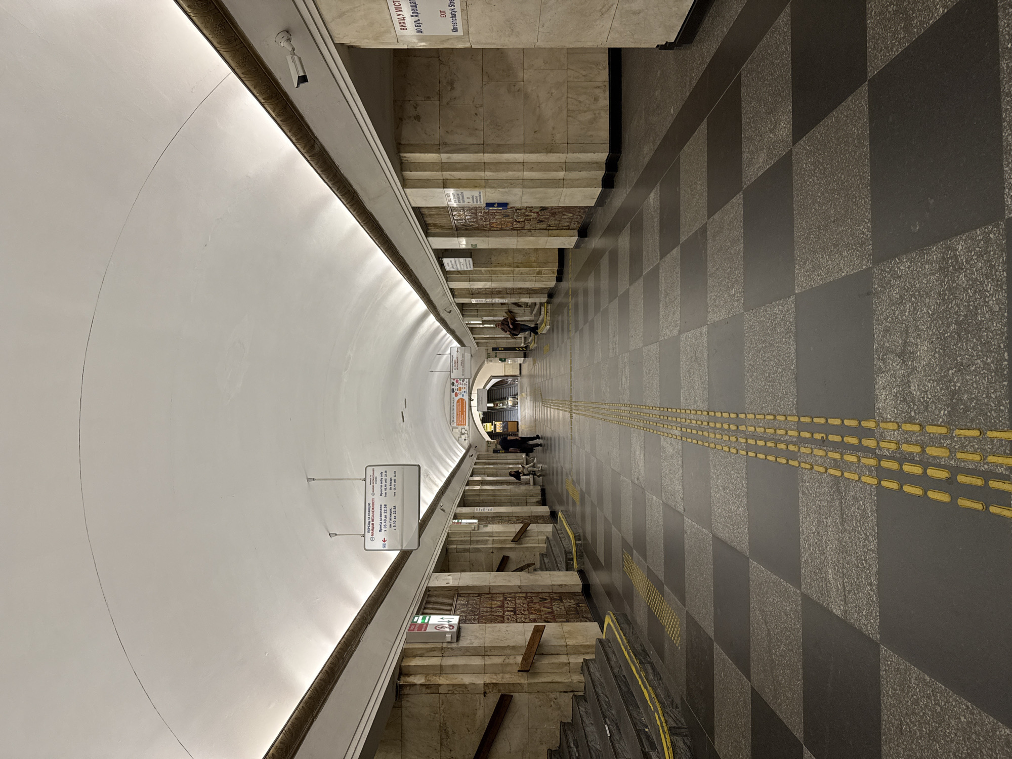 The image shows a wide, empty metro station platform with a high, arched ceiling. The floor is tiled in a checkerboard pattern of dark gray and lighter gray squares. Yellow tactile paving lines run down the center of the platform, indicating a safe walking path. On either side of the platform are stone-clad walls with staircases leading up or down. Several signs are mounted on the walls, some with Cyrillic text. There are a few people present: one person sitting on a bench on the left, and two people standing further down the platform on the right. A security camera is visible mounted on the ceiling near the right wall. The overall impression is one of quiet and order, with clean lines and a symmetrical design.
