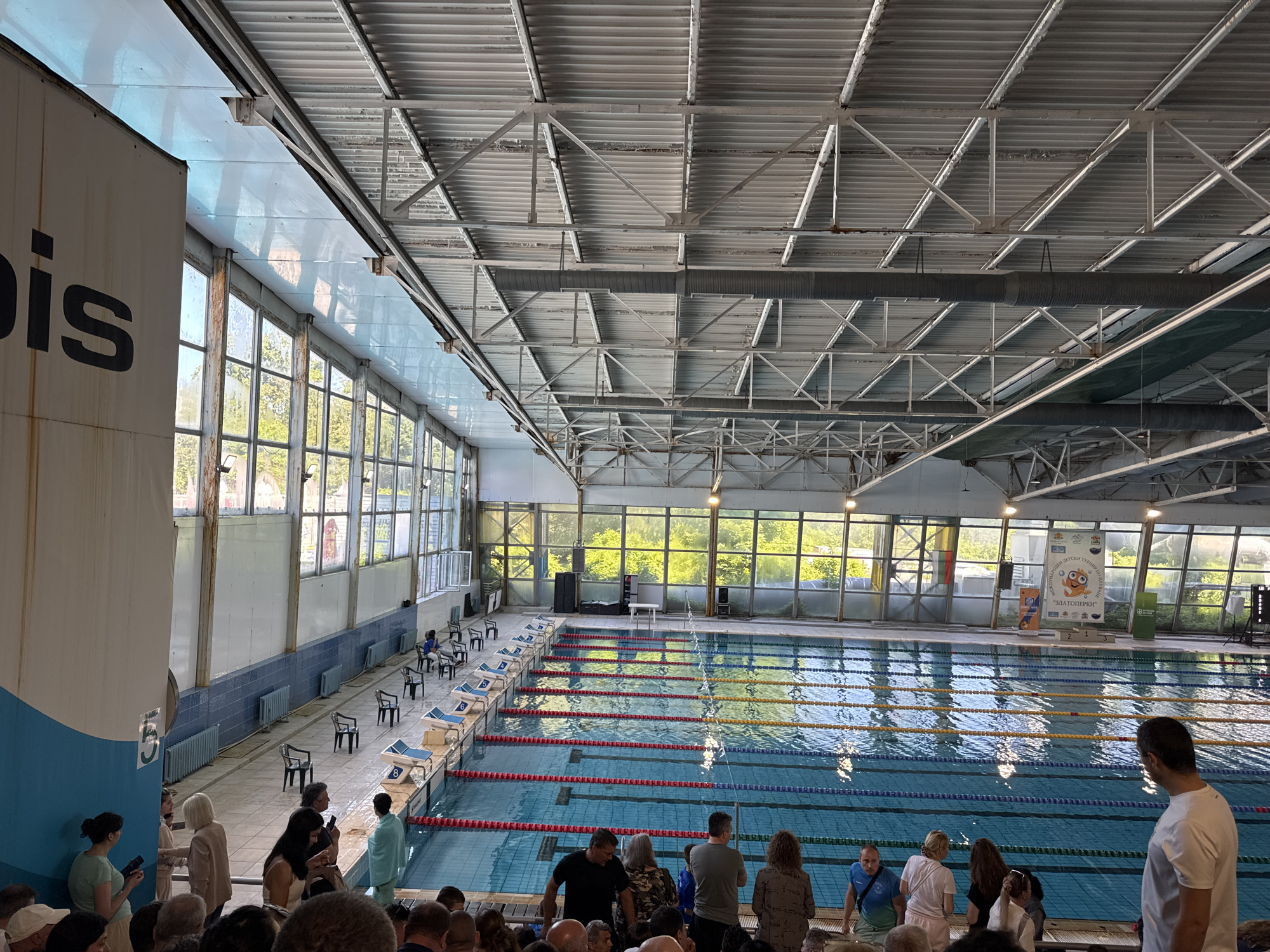 A high-angle shot of a competition swimming pool. The pool is rectangular with multiple lanes separated by lane markers. Starting blocks are positioned at the end of each lane. The pool is filled with water with reflections of light visible on the surface.
Along the left side of the pool, there are rows of windows. A partially obscured sign is visible on the left side of the frame. The windows offer a view of greenery outside. The building has a high ceiling with exposed metal support structures and ductwork.
People are standing and sitting around the pool, many appearing to be spectators. The crowd appears dense, with people of various ages and ethnicities.
Located in Sofia, Bulgaria.