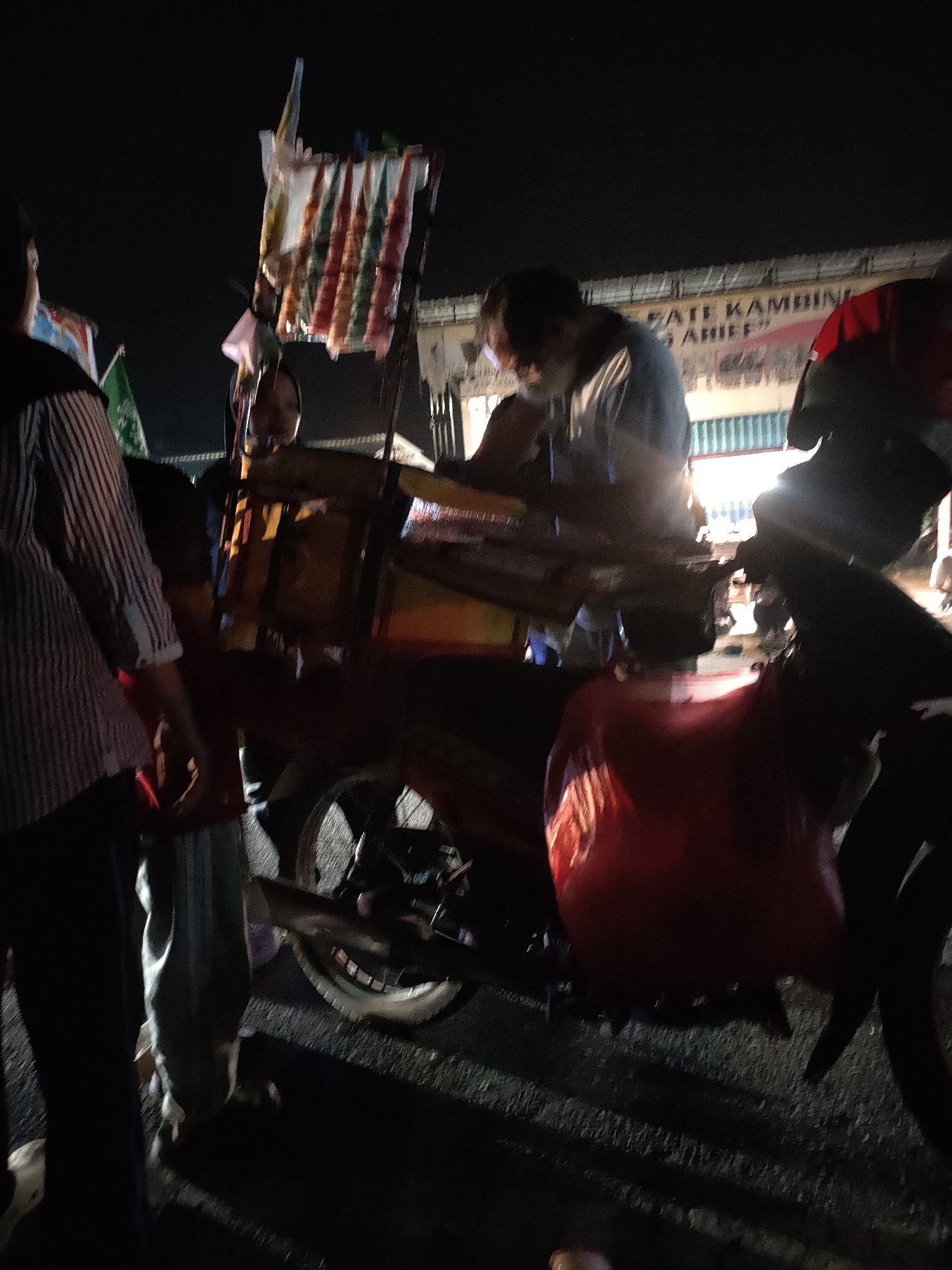 The image shows a dimly lit street scene in Kanigoro, Indonesia at night. A motorcycle with a sidecar is parked, with a vendor attending to it. The sidecar is laden with various items, including what appear to be colorful snacks or food items hanging from a display. A sign above the motorcycle reads "SATE KAMBING ARIE". Several people are present, some blurred due to movement or low light. One individual, possibly the vendor, is seen leaning over the sidecar, appearing to be engaged in a transaction. Another person, wearing a striped shirt, is visible on the left side of the frame. The background suggests a building with some lighting. The ground is paved, and some grass is visible. The overall atmosphere is that of a street food stall operating at night.