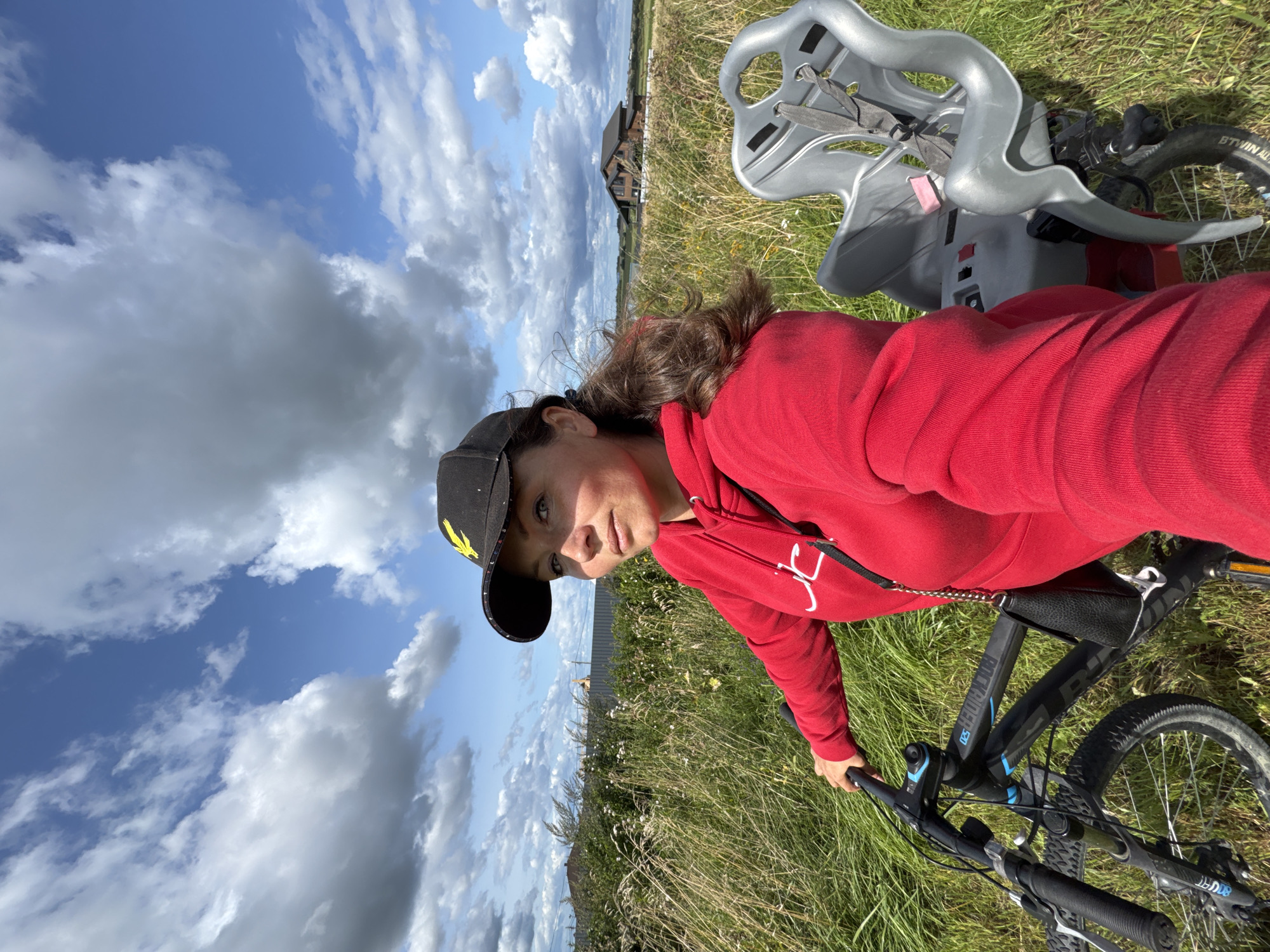 A woman wearing a red hoodie and a black baseball cap with a yellow eagle logo is posing for a selfie outdoors. She is holding onto the handlebars of a bicycle, which has a child seat attached to the back. The bicycle is parked on a grassy area with tall dry grass and some white flowers. In the background, there is a wooden house with a dark roof, a fence, and a cloudy blue sky. The scene appears to be in a rural or suburban area, possibly on the outskirts of Kempelevo, Russia.