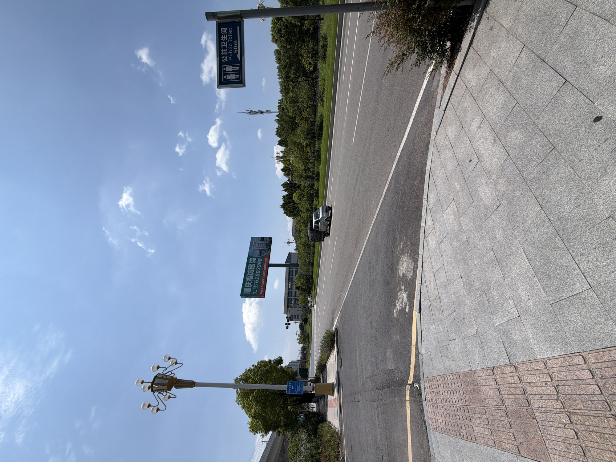 The image depicts a street scene in Jiangjin, China, on a sunny day with scattered clouds. A wide, paved road stretches into the distance, with a white lane divider. On the right side of the road, a blue sign indicates "Public Toilet 50m" with pictograms for men and women. Further down the road, a white van is visible.
To the left of the road is a sidewalk paved with gray and reddish-brown tiles, with a textured tactile paving section for visually impaired individuals. A lamppost with multiple lights stands on the sidewalk. Behind the lamppost, a billboard advertises "Chongqing Jiangjin Hospital" with a phone number. Trees and greenery line the right side of the road and the background. In the distance, a cell tower is visible. The overall impression is a well-maintained urban environment.