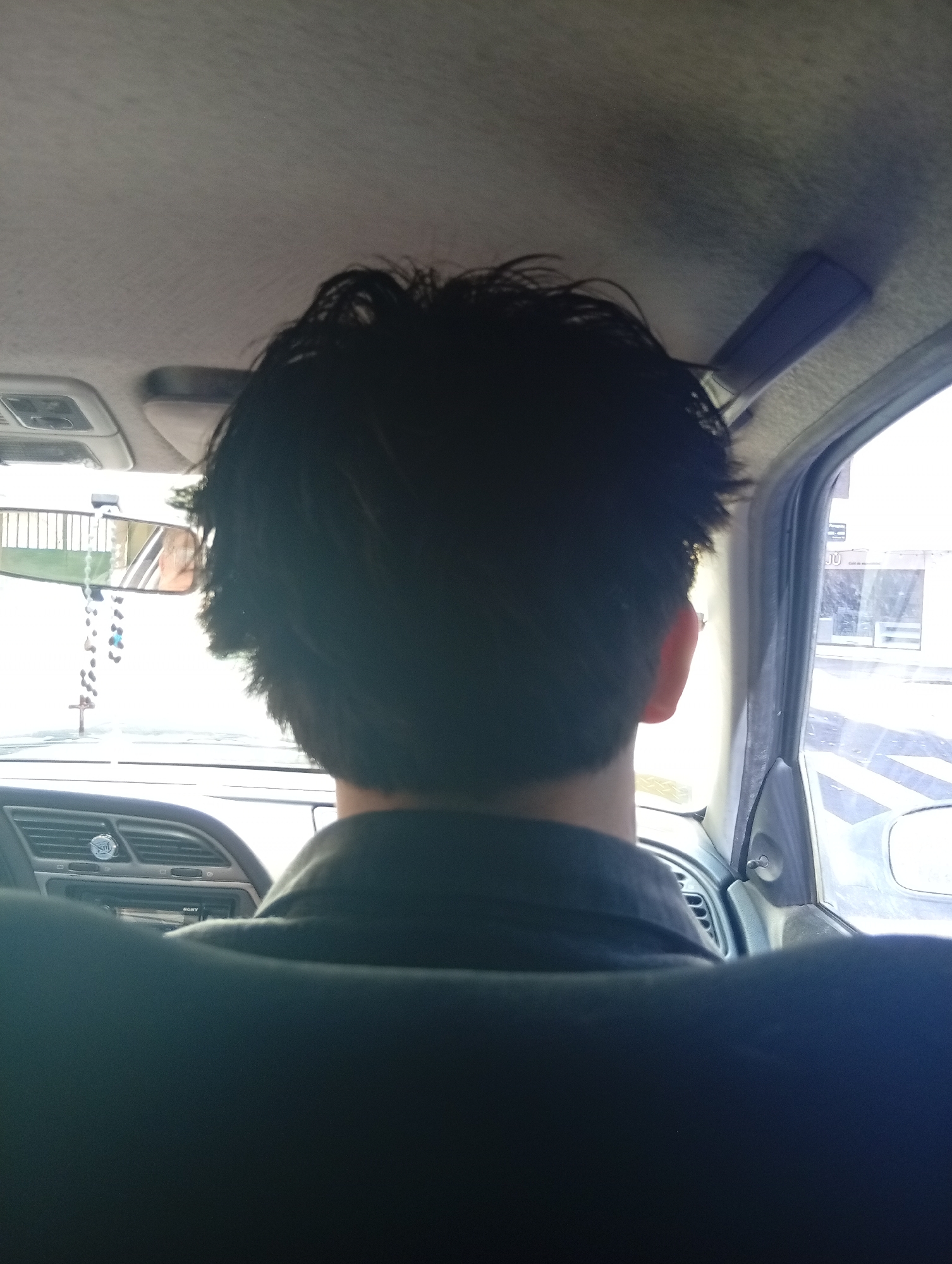 The image depicts the back of a person's head, with dark, messy hair, inside a car. The person is wearing a dark shirt. The interior of the car is visible, including the rearview mirror with a beaded rosary hanging from it, the dashboard, the ceiling, and a side window. Through the side window, a street scene can be seen, including a building with the letters "JU" visible. The scene is likely taking place in Buenos Aires, Argentina, due to the street signs and architecture visible through the window. The car is in motion, though it is unclear if the person is the driver or a passenger.