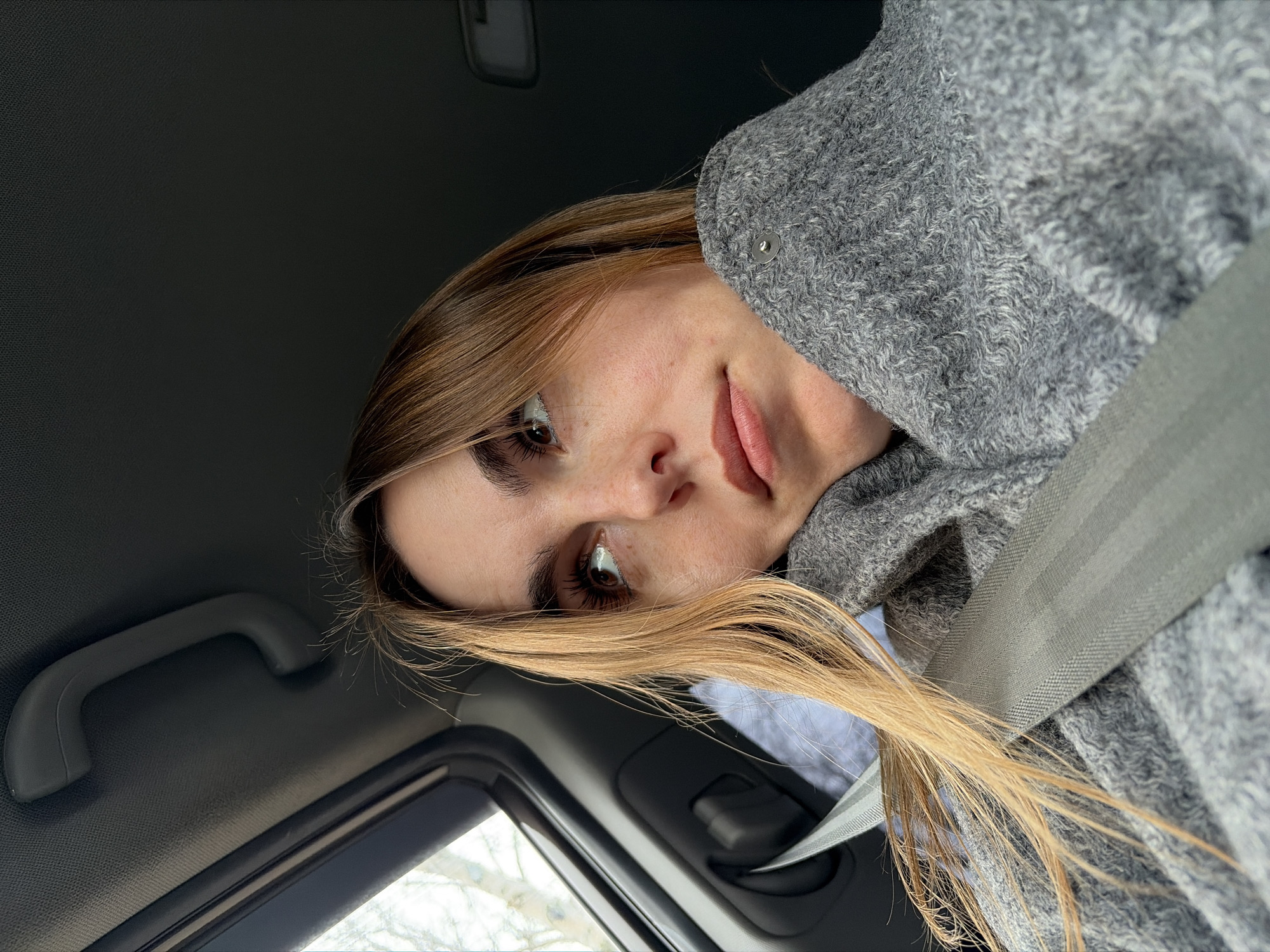 The image shows a young woman wearing a gray sweater and a seatbelt inside a car. She has light brown hair and is looking towards the camera. The interior of the car is visible, including the dashboard, a window, and a grab handle. The image provides no information about the location in Izhevsk, Russia.