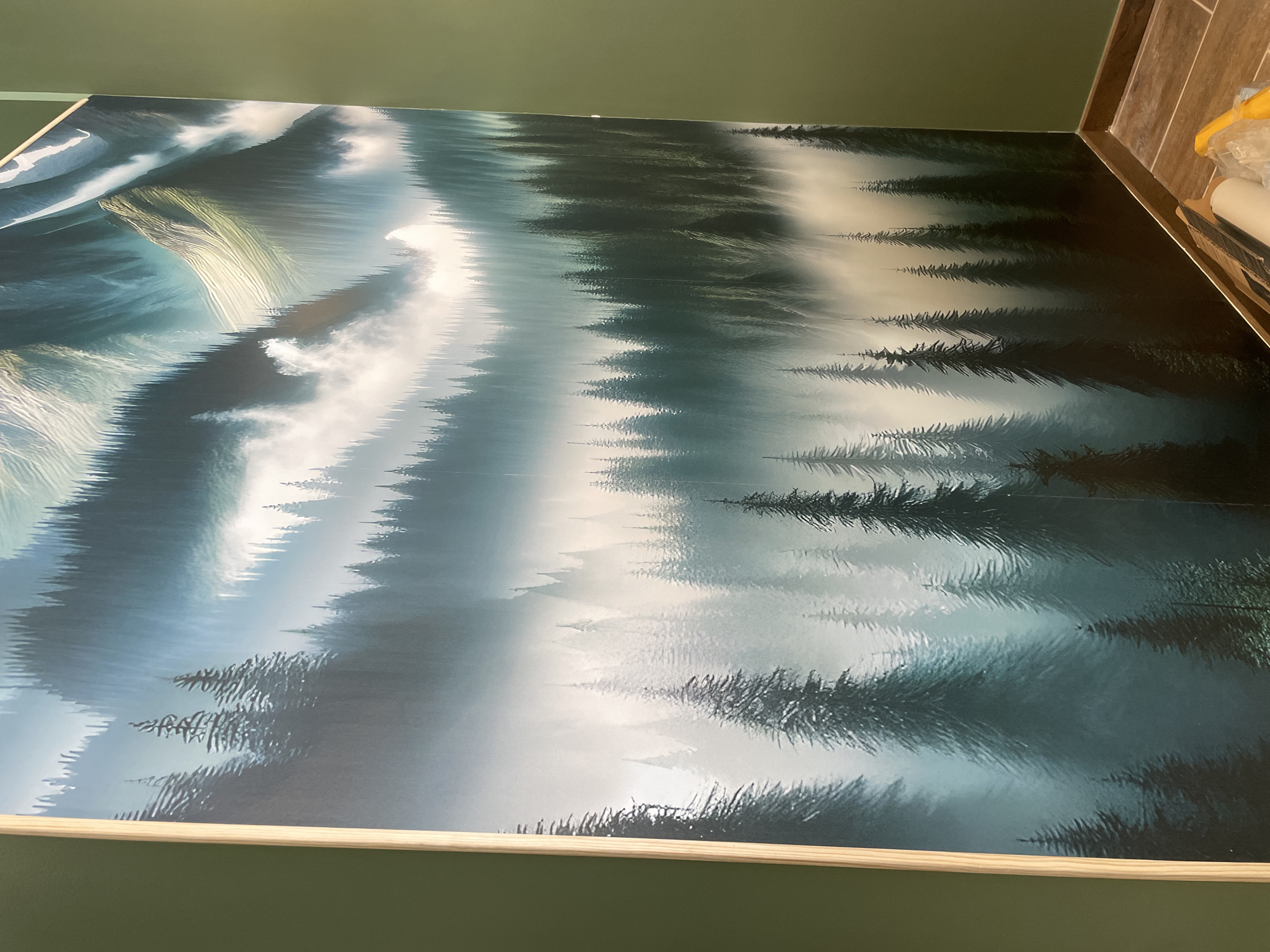 The image displays a large, vibrant mural of a misty forest with majestic mountains in the background. The mural is mounted on a green wall. To the right of the mural, there is a corner where the green wall meets another green wall, and a wooden baseboard runs along the bottom. On the floor in this corner, there are tools likely used for installing the mural, including rolls of material, a scraper, and a yellow object that may be a bag or tarp. The scene is devoid of people. The mural's artistic style is somewhat stylized and evocative, with a focus on atmospheric lighting and deep greens and blues. The overall impression is that of a new installation in progress or recently completed within an interior space. There is no indication or information linking this image to the city of Meaux, France.