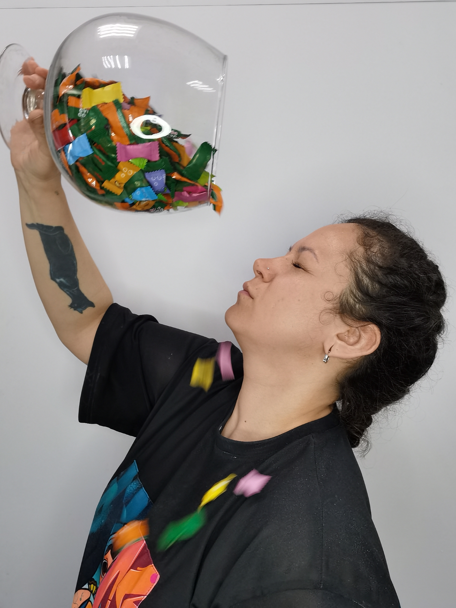 A person is standing against a white wall, holding a large glass bowl filled with colorful candy wrappers above their head and tilted toward their face. Some wrappers are falling from the bowl. The person has dark curly hair pulled back, a nose piercing, and is wearing a black t-shirt with a colorful graphic design on the front. They have a tattoo on their left upper arm. The city and country of Chaikovsky, Russia are not evident in the image.