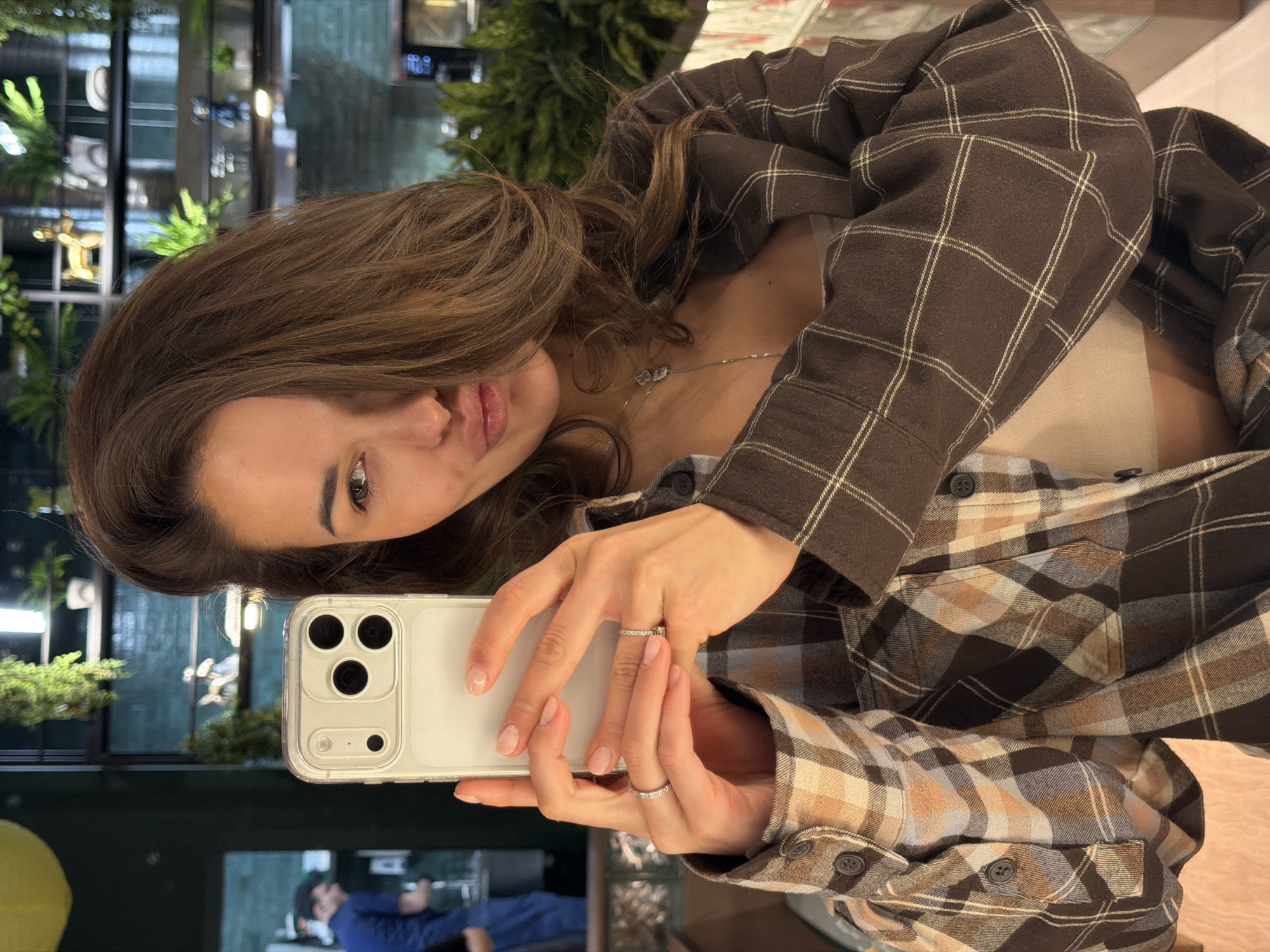 A young woman is captured in a selfie taken indoors, likely within a modern cafe or restaurant in Krasnoyarsk, Russia. She is shown from the chest up, with her head slightly tilted, looking directly towards the phone's camera. Her long, brown hair cascades around her shoulders.
She is wearing a brown plaid flannel shirt with white and lighter-toned grid patterns over a light, possibly beige or cream, undershirt. A delicate silver necklace with two small charms, possibly hearts, adorns her neck. In her left hand, she holds a light-colored smartphone, likely an iPhone given the camera lens configuration, encased in a clear protective cover. Two silver rings, one a simple band and the other featuring small embedded stones, are visible on her left ring finger. Her fingernails have a natural, neatly manicured appearance.
The background reveals a contemporary interior space characterized by large glass panels or windows, which reflect parts of the room, including a glimpse of another person in a blue top in the lower-left reflection. Lush green plants, some hanging, are strategically placed, enhancing the modern aesthetic. Artificial lighting fixtures are also discernible in the background. The scene suggests a casual, everyday moment. No visible text is present in the image.