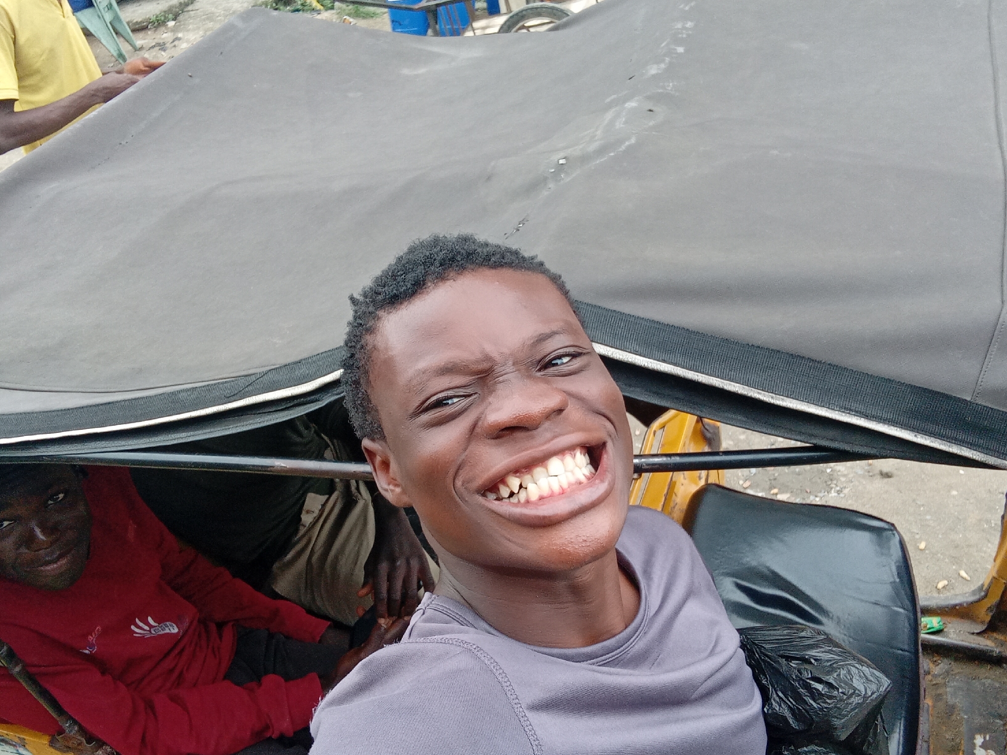 In a city setting of Unknown, Unknown, a young Black male in a grey t-shirt is smiling widely, holding up his phone to take a selfie inside what appears to be a motorized tricycle. He is positioned in the foreground, with his face prominently displayed and his mouth open in a broad grin, revealing his teeth. Behind him, another young Black male wearing a red long-sleeved shirt is visible, seated and looking towards the camera. A portion of a dark-colored tarp or canopy is above them, with metal bars supporting its structure. The environment appears to be outdoors, with a rough, paved surface visible in the background. A black plastic bag sits on a black seat to the right of the frame. The scene suggests a candid moment captured during transportation.