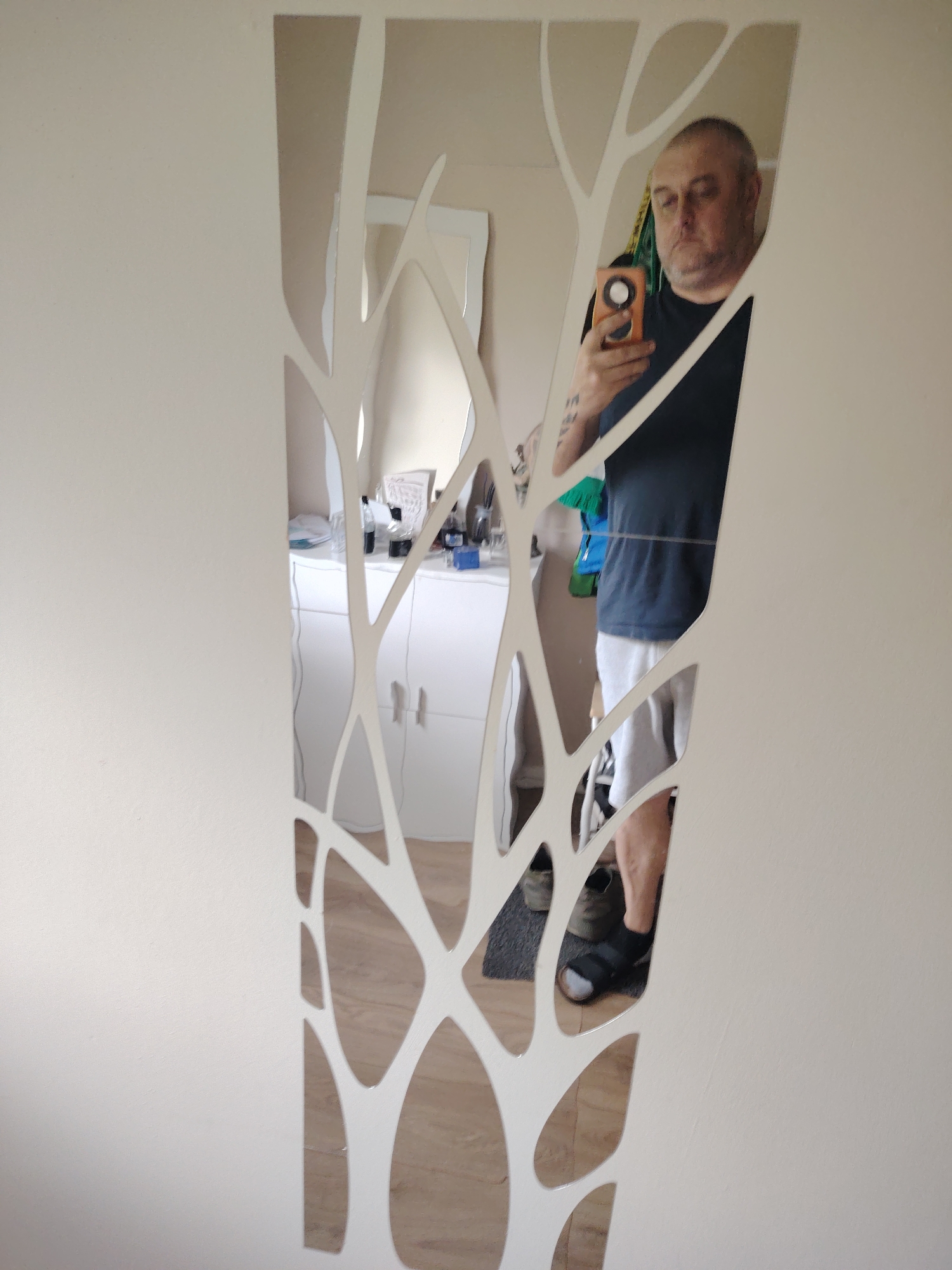 The scene depicts an interior view in Huntingdonshire, United Kingdom, captured via a selfie reflected in a decorative wall mirror. An adult male, with a bald crown, short grey hair on the sides, and a full face, is centered in the reflection. He wears a dark short-sleeved t-shirt and light-colored shorts. On his left forearm, a tattoo is partially visible. He holds an orange rectangular smartphone with a circular attachment in his right hand, pointed towards himself, indicating he is taking the photograph. His gaze is directed downwards, possibly at the phone's screen.
The mirror itself is a vertical, narrow decorative piece mounted on a plain, light-colored wall. Its design features irregular, branching, and curvilinear white frames surrounding multiple reflective sections, resembling abstract tree branches or roots.
Reflected in the mirror behind the man is a room interior. To the right of the man's reflection, a white cabinet or dresser with multiple doors or drawers is visible, topped with various items, including clear bottles, a blue container, and some white fabric. Higher up, a white-framed mirror or picture frame is partially reflected. To the left of the man's reflection, glimpses of additional furniture and possibly draped fabric (yellow and green) are seen. The floor visible in the reflection is light-colored wood laminate, with a dark mat or rug positioned directly beneath the man's feet, which are clad in black sandals worn over white socks or a white sandal band.
