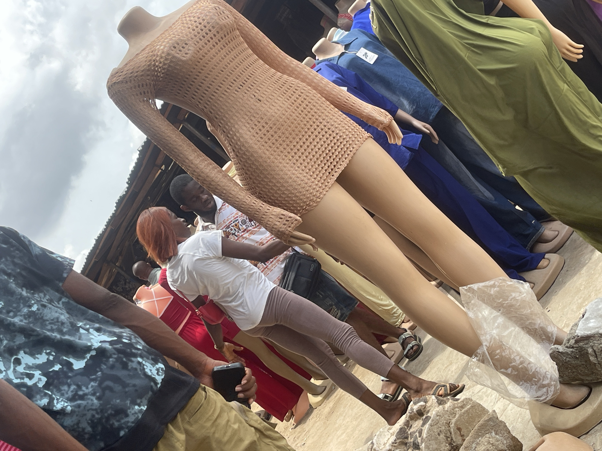 The image shows a street scene in an unidentified city. Several mannequins dressed in various types of clothing are positioned in a row. The first mannequin is wearing a tan, knitted dress, and another is dressed in a blue denim shirt and dark blue pants. A green dress covers another mannequin. Several people are visible in the background. One individual is wearing a white shirt and gray leggings, and another is wearing a red dress. One person is looking at the mannequin dressed in the tan, knitted dress. The ground is paved and rough, with some rocks and a plastic bag near the mannequins' feet. The sky is overcast, suggesting it might be a cloudy day.
