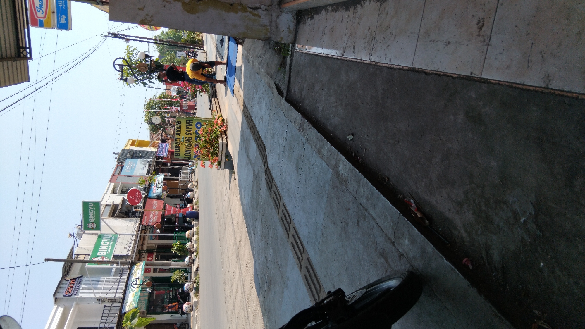 The image depicts a street scene in Batang, Indonesia, on a clear, sunny day. The foreground is dominated by a concrete sidewalk with tactile paving, suggesting a public walkway. To the left, a portion of a motorcycle is visible.
The middle ground of the image reveals a street with a few parked motorcycles and several shops lining the opposite side. These shops have various signage, including "BINGYU" (repeatedly), "K NOFONO," and "LaScion." There is a green sign in front of the shops that reads "BUBUR AYAM LONTONG SAYUR MOTO." Two men are present on the sidewalk near this sign. One man, wearing a yellow shirt, is bending over, possibly engaged in an activity on a blue mat. Another man, in a black shirt, stands nearby, looking towards the street. The environment appears to be an urban commercial area.
The background shows a clear blue sky, power lines, and more buildings and structures, indicating a typical streetscape. The overall impression is of a moderately busy commercial street.