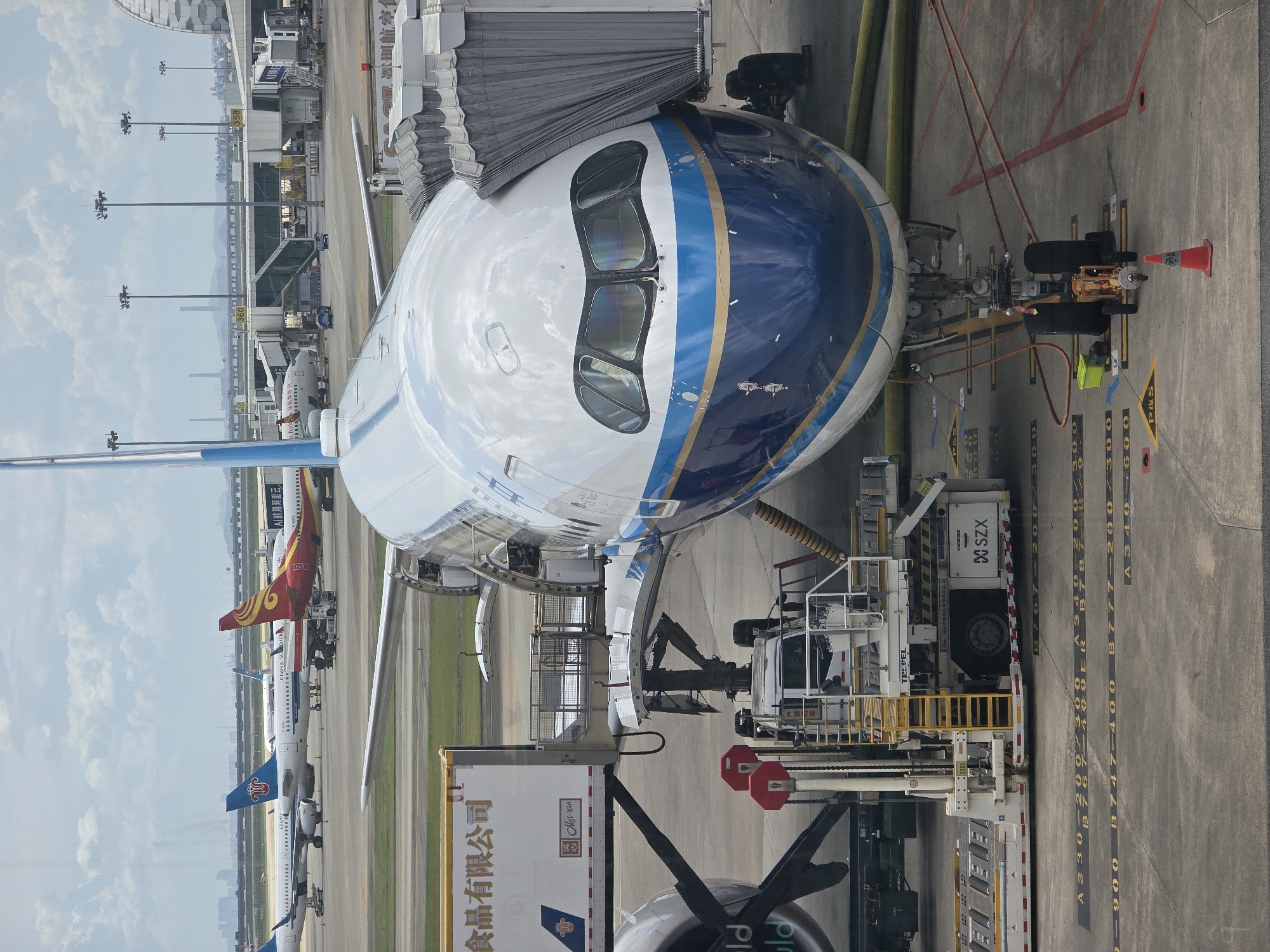 The image shows an aerial view of an airport tarmac in Bao'an District, China, on a partly cloudy day. A large passenger airplane, painted white with blue and gold stripes, is parked at a gate, connected to a jet bridge. In the foreground, a baggage loading cart is positioned near the aircraft's cargo hold. Other airplanes are visible in the background, along with airport buildings and structures, including jet bridges and control towers. The tarmac is marked with yellow lines and symbols indicating aircraft parking positions and taxiways. The overall scene depicts a busy airport environment with aircraft undergoing loading and unloading procedures.