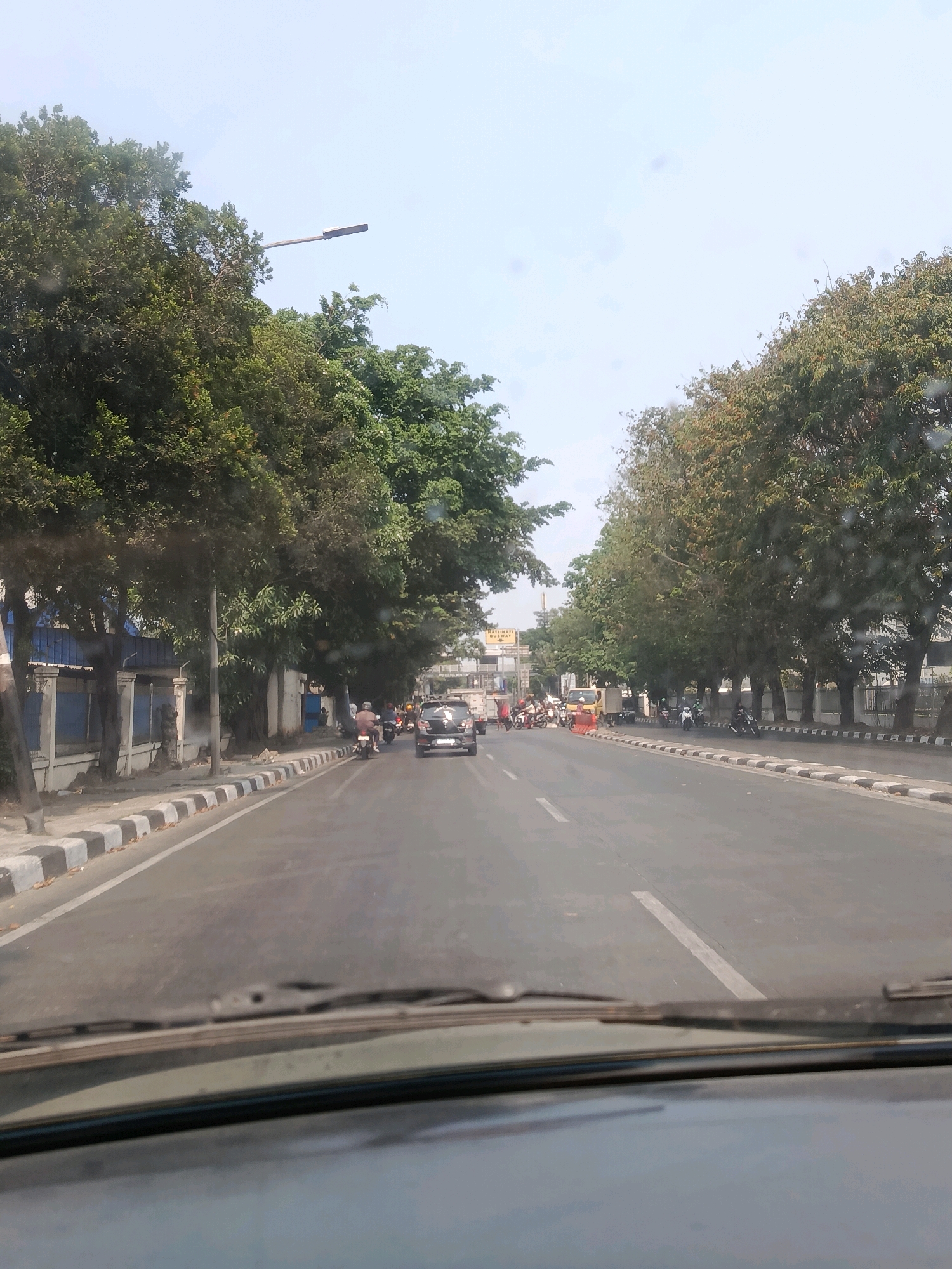 The image depicts a street scene in East Jakarta, Indonesia, viewed from the interior of a vehicle. A two-lane road stretches into the distance, bordered by a sidewalk and lined with trees on both sides. Several vehicles, including cars and motorcycles, are present on the road, with some appearing to be in motion. A streetlight is visible above the trees on the left side of the image. The environment appears to be a sunny day with a clear sky. Buildings and fences are visible along the roadside on the left. The perspective suggests the photograph was taken while driving.