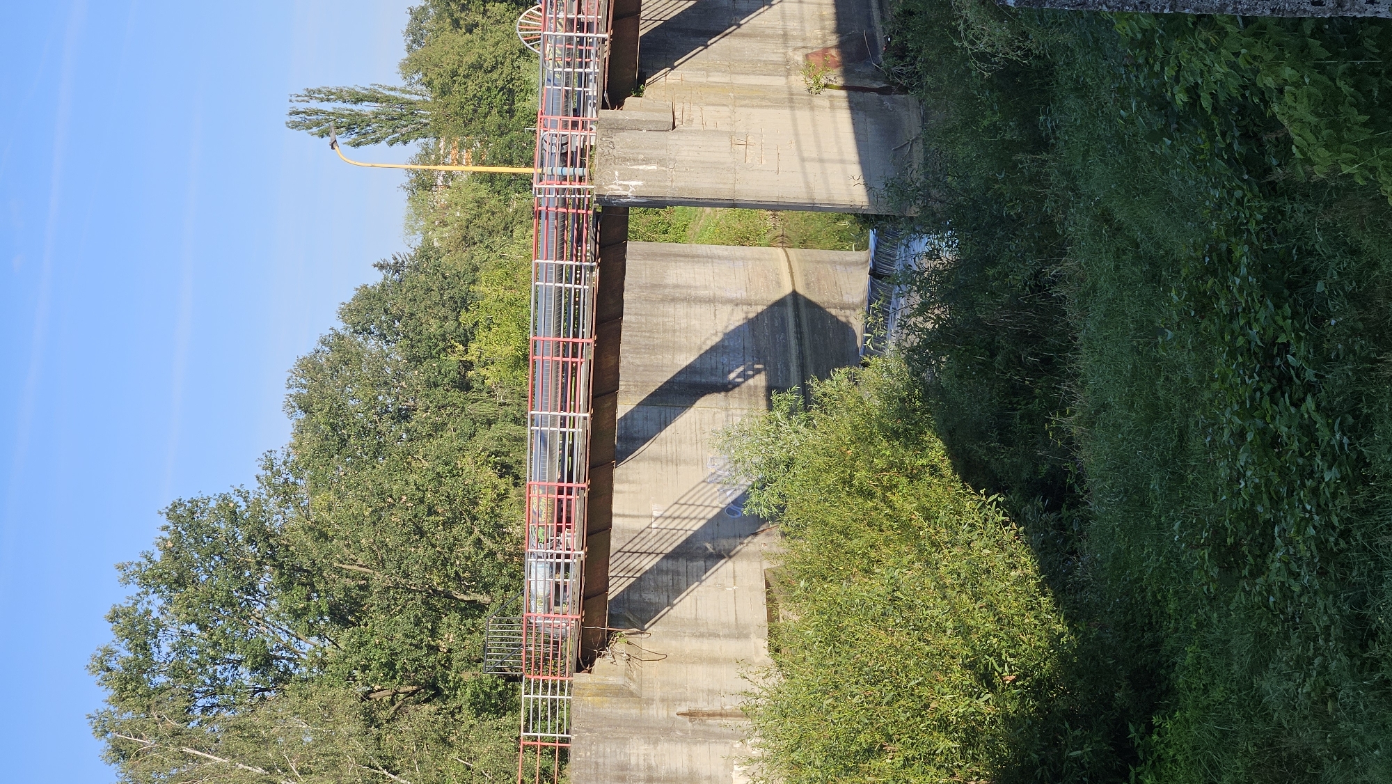 The image depicts a concrete bridge spanning a small waterway, likely a canal or a narrow river, in Prešov, Slovakia. The bridge has a railing with red and white painted poles and horizontal metal bars. A large pipe runs along the length of the bridge. Concrete abutments support the bridge, and the waterway below has a small waterfall or weir. Lush green vegetation, including trees and bushes, surrounds the scene, with a clear blue sky overhead. A lamppost is visible in the background to the right. The lighting suggests it is daytime, with shadows cast by the bridge structure. There are no people or animals visible in the image.