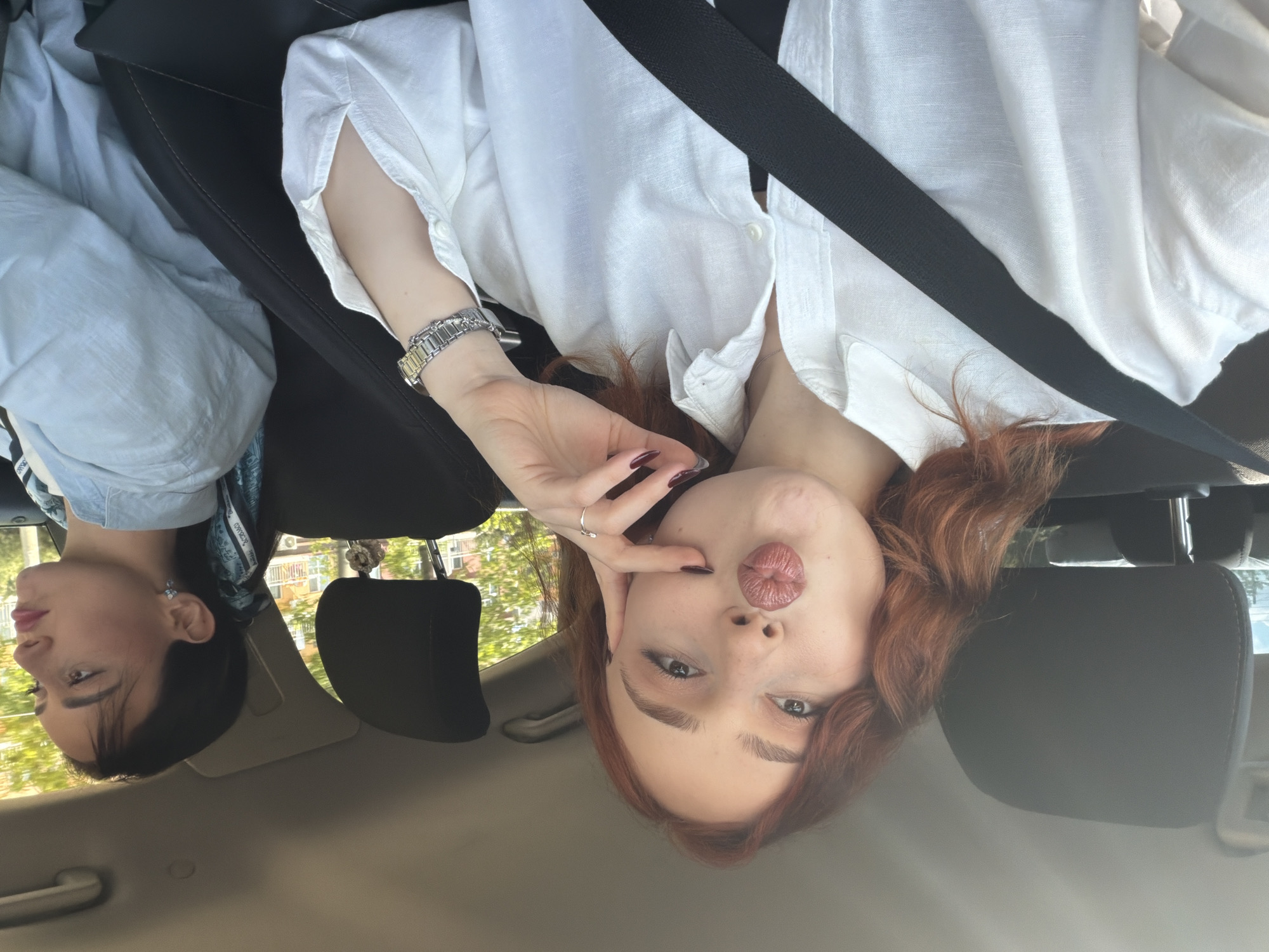 The image captures two women inside a car, likely a selfie or casual photo taken during the day. The primary subject, on the right side of the frame, is a woman with reddish-brown, wavy hair. She has a light complexion and is wearing a white collared shirt. A silver watch with a metallic strap is visible on her left wrist, and her fingernails are painted with dark polish, possibly burgundy. She is looking directly at the camera with wide eyes and her lips are pursed in a "kissy face" or "duck face" gesture, with one hand near her cheek. A black seatbelt diagonally crosses her chest.
To her left, partially visible in the background, is another woman with dark hair and bangs. She has a light complexion and is wearing a light blue top. A patterned scarf, featuring light blue and white designs with visible text including "DIOR", is tied around her neck. She is looking away from the camera, seemingly towards the left (viewer's right).
The setting is the interior of a car, with black leather seats and headrests visible. Through the car windows, trees and residential buildings can be seen, indicating an outdoor, urban, or suburban environment during daylight hours. The natural light suggests a sunny day. The overall impression is a casual moment captured while traveling in Tbilisi, Georgia.