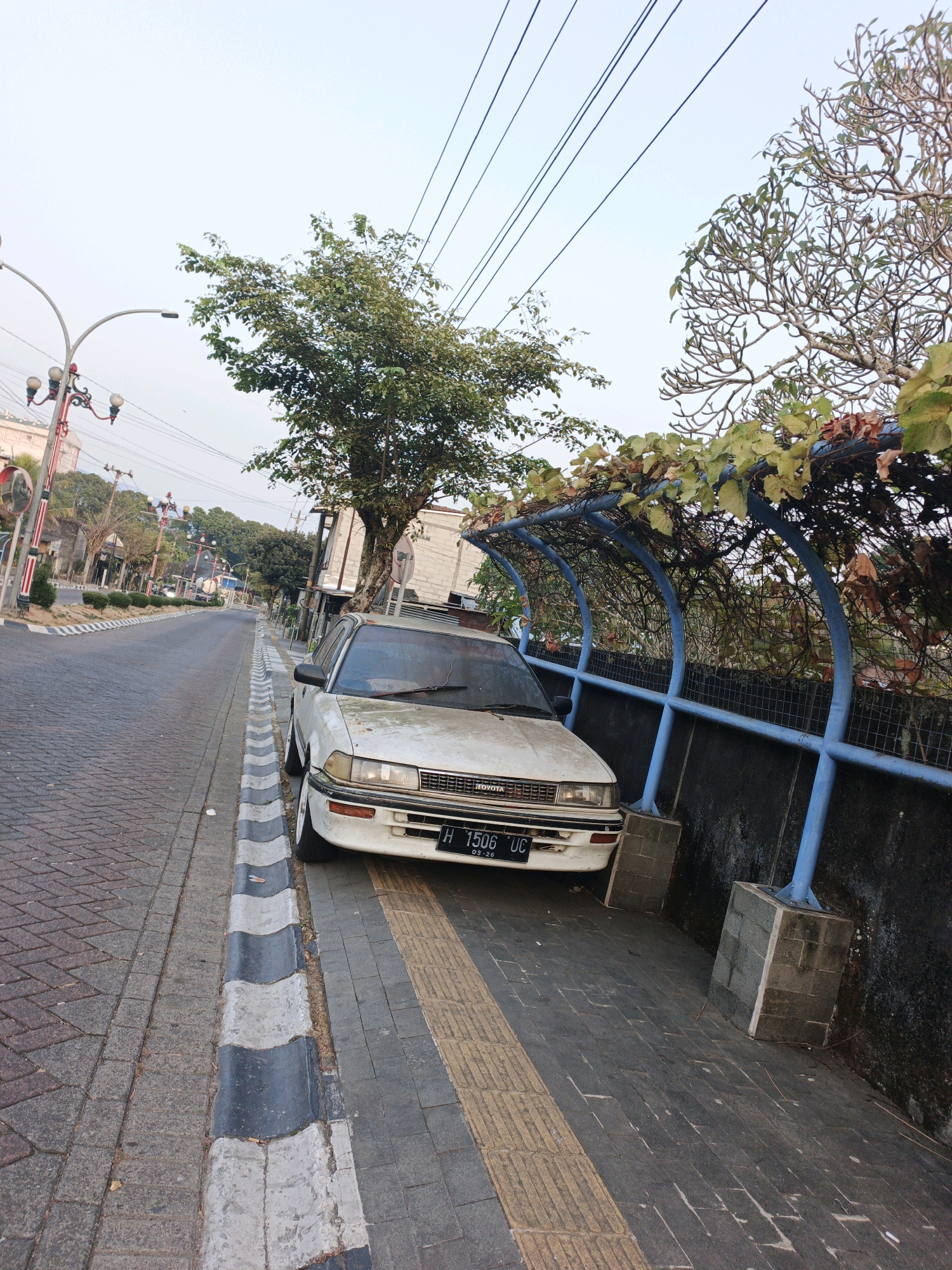 A white Toyota sedan is parked on a paved sidewalk next to a street in an urban environment. The car is facing left and is angled slightly towards the street. The license plate reads "H 1506 UC". The car appears to be old and somewhat weathered. The sidewalk is made of interlocking bricks and has a yellow tactile paving strip. A low, grey, undulating curb separates the sidewalk from the street. The street is paved with similar interlocking bricks. In the background, there are trees, buildings, and utility poles with numerous wires overhead. A blue metal structure, possibly a bus stop shelter or a decorative archway, covered in vines, is located behind the car. The sky is overcast. The scene suggests a typical street view in a city.