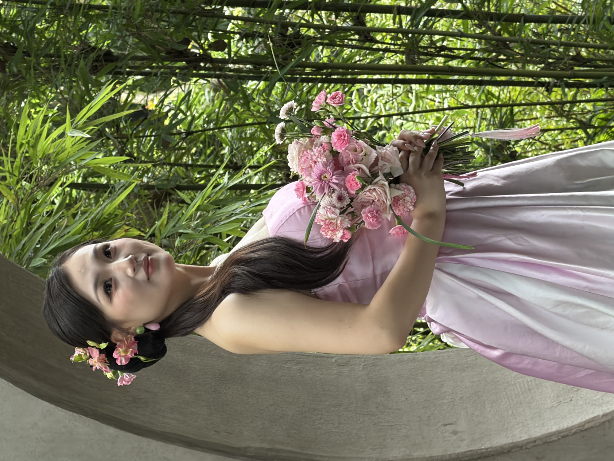 A young East Asian woman with dark hair styled in a bun adorned with pink flowers and teardrop-shaped earrings poses in a strapless light pink dress. She is holding a bouquet of pink and white flowers. The woman is positioned in front of a circular stone archway, with a dense bamboo forest visible in the background. The scene appears to be outdoors, possibly in a garden or park. The overall mood is serene and elegant. There is no identifiable location information in the image that would place it in Yuzhong District, China.