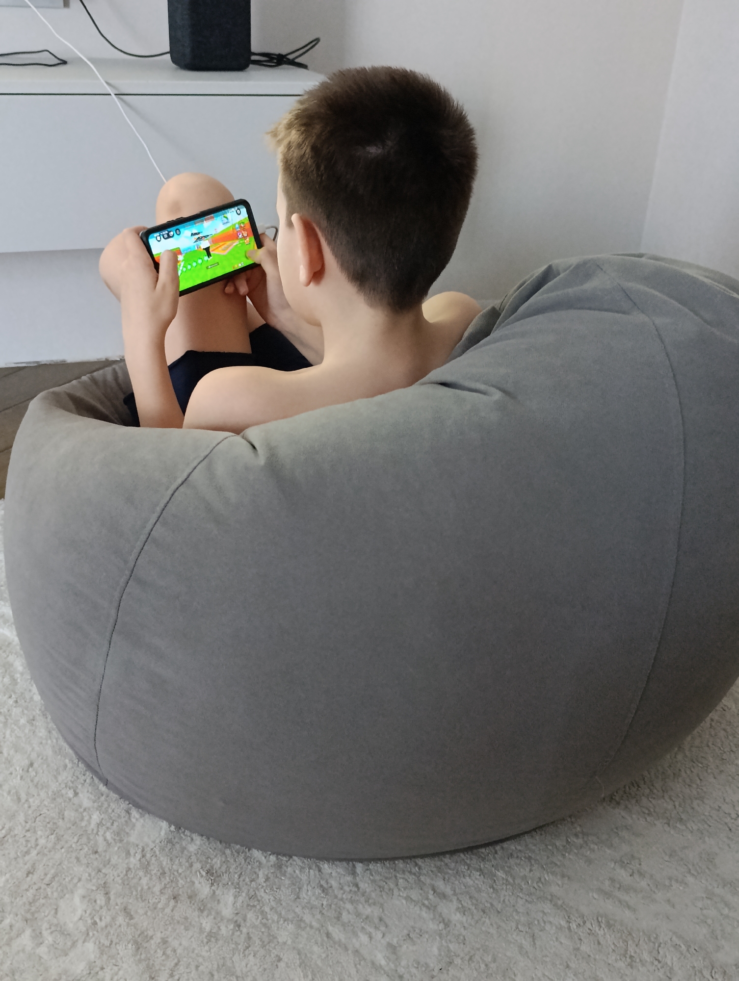 A young boy sits in a grey beanbag chair and plays a game on a black smartphone. The boy has short, light brown hair, and his back is bare. He appears to be in a room with white walls and a white shag carpet. In the background, there is a white cabinet and a black speaker. The location is Chaikovsky, Russia.