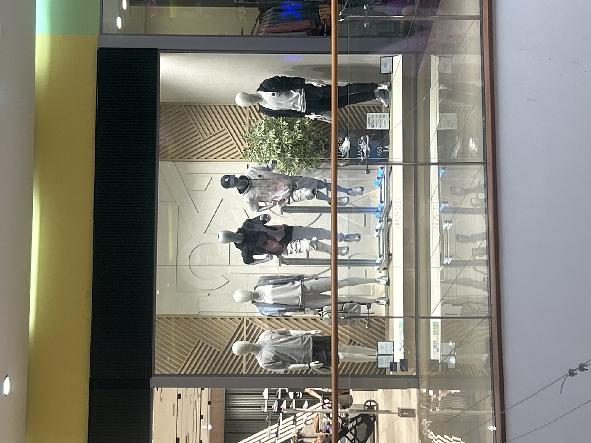 This image captures a retail store display through a large glass window, likely within a shopping mall. The scene is viewed from an angle, with the store's interior well-lit by artificial light.
The primary subjects are five male mannequins arranged vertically within the display. Each mannequin is dressed in contemporary athletic or casual clothing.
1. The topmost mannequin wears a black jacket over a white top, black pants, and sneakers.
2. Below it, another mannequin sports a light-colored, possibly patterned, t-shirt with light shorts and a black cap. A small green plant is positioned behind this mannequin.
3. The third mannequin wears a dark long-sleeved top with light pants and sneakers.
4. The fourth mannequin is dressed in a light grey jacket over a white t-shirt, light pants, and sneakers.
5. The bottommost mannequin features a grey t-shirt, black shorts, and sneakers.
The display's background consists of white walls with subtle geometric patterns and textured wooden panels featuring diagonal slatted designs. Various display stands and signs are scattered throughout.
The large glass window creates numerous reflections of the surrounding mall environment, including what appear to be escalators or staircases, overhead lighting fixtures, and indistinct shapes of other parts of the building. These reflections partially obscure some details within the store display.
The brand name "LLAGGI" is prominently visible multiple times on bags and display materials inside the window. Several smaller white signs with unreadable text are also present.
The store's exterior is framed by a dark black vertical panel on the left, adjacent to a yellow and light green painted pillar. The right side of the window has a brown frame next to a plain white wall. The overall setting suggests a modern retail space in a shopping center in Pleven, Bulgaria.