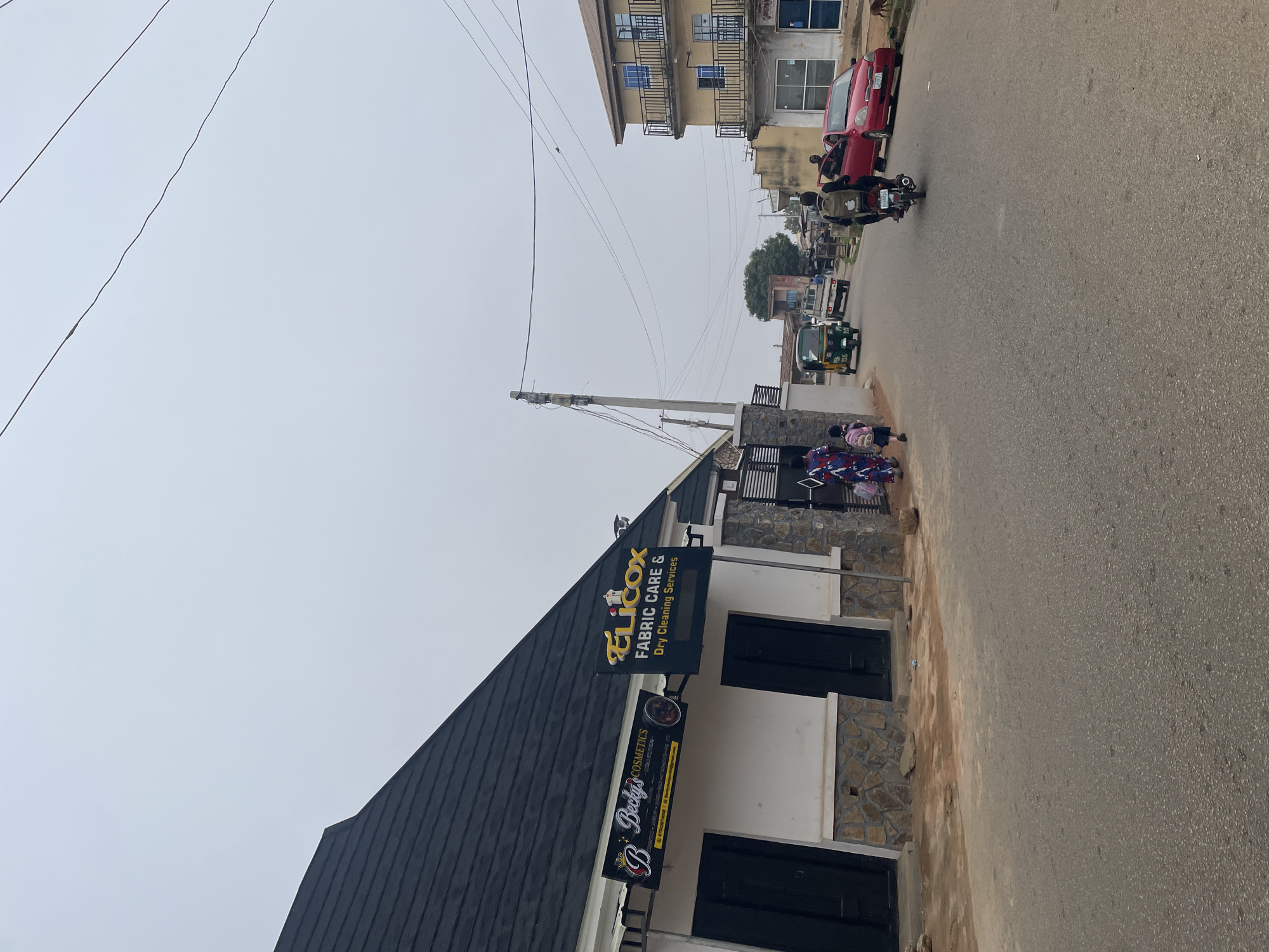 A street scene in Jos, Nigeria, under an overcast sky, features a commercial building with two businesses. The building's left side displays a sign for "Becky's Cosmetics" and on the right, a sign for "Elicox Fabric Care & Dry Cleaning Services." The facade is a mix of white paint and stone cladding. Adjacent to the building, a black metal gate is visible.
In the street, several vehicles are present. A red car is parked on the right side, and a motorcycle with a rider wearing a backpack is moving away from the camera. A tricycle, also known as a Keke Napep, is traveling down the street, followed by a white pickup truck.
On the left side of the street, near the commercial building, two young females are walking away from the camera, one carrying a pink backpack. Further down the street, other people are visible near buildings. The environment appears to be a developing urban area with a mix of residential and commercial structures, overhead utility wires, and a paved road surface.