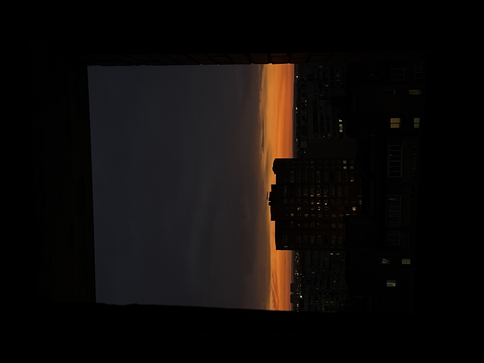 The image depicts an urban landscape at twilight or sunset, viewed from an interior perspective looking out through a dark, indistinct rectangular opening, likely a window or doorway. The interior framing elements on the left, right, and top are solid black, contrasting sharply with the illuminated exterior.
The sky occupies the upper two-thirds of the visible outdoor scene. The uppermost portion is a dark, desaturated gray-blue, transitioning downwards into a prominent horizontal band of vibrant orange and yellow light indicative of a sunset, with subtle cloud formations visible within the upper gray area and at the transition to orange. A faint, distant line of city lights is visible along the horizon, just below the brightest orange.
In the midground, a large, dark multi-story residential building stands prominently in silhouette against the bright orange sky, slightly right of center. Numerous windows on this building emit yellow and white light from within. To its left, other, less distinct, dark building silhouettes are visible. Further down and to the left, two prominent, lighter-toned rectangular structures with flat tops are discernible, possibly industrial or architectural features.
The lower portion of the visible exterior scene consists of a dark cityscape, with various building outlines merging into the shadows. Some scattered window lights are visible in these darker structures, particularly in the lower midground. No individuals are visible anywhere in the scene. The overall lighting creates a high-contrast environment, with deep shadows dominating the foreground and framing, and bright warm hues in the sky.