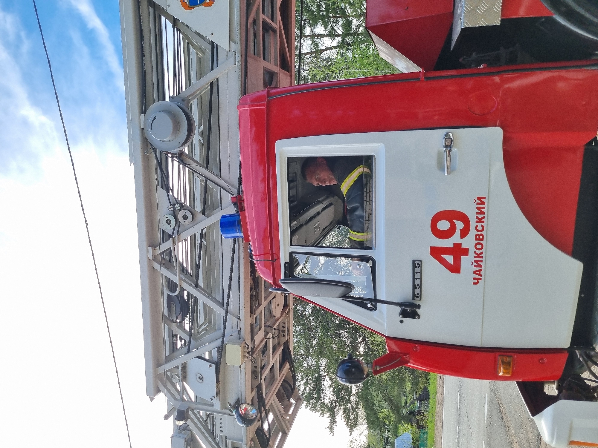 The image shows a fire truck with its ladder extended upwards. The truck is red and white and has the number "49" and the word "Чайковский" written on its side, indicating its location in the city of Chaikovsky, Russia. A firefighter is visible inside the cab of the truck. The ladder is made of a white metal structure and extends towards the sky, where some clouds are visible. The environment around the truck includes trees and other greenery. The image appears to be taken outdoors on a daytime, suggesting that the scene may be related to a fire emergency or training.