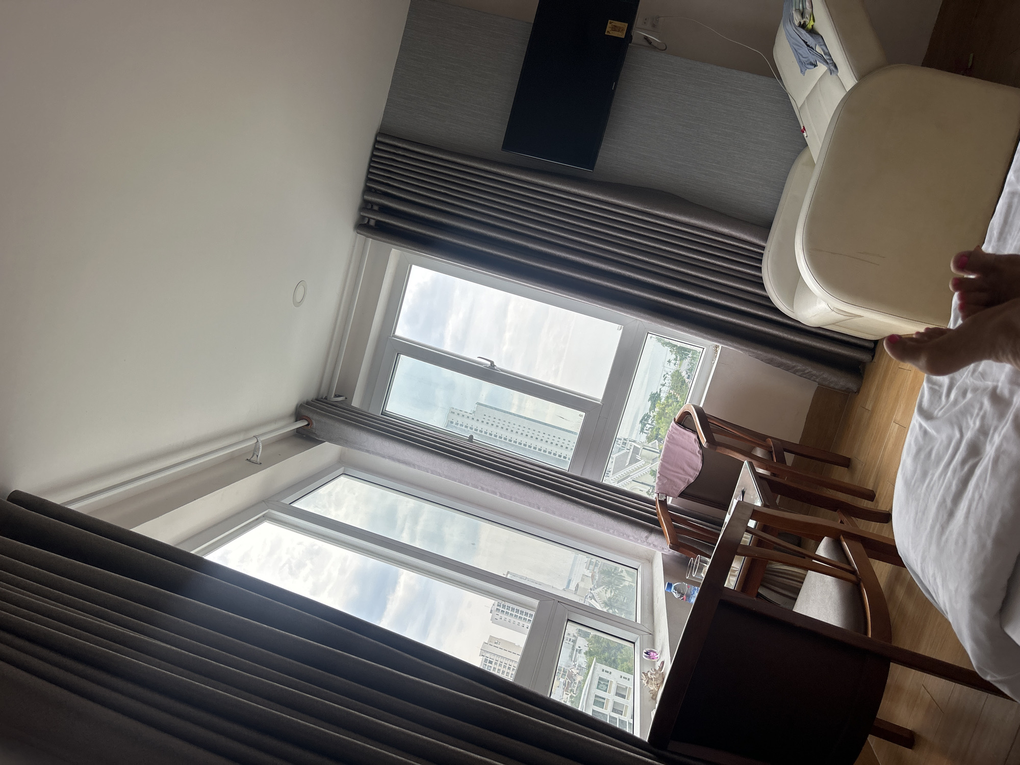 The scene is an interior view of a room in what appears to be a high-rise building, likely a hotel or apartment, in Bac Nha Trang Ward, Vietnam. Two large windows are visible, each covered with gray curtains that are partially drawn. Through the windows, a cityscape is discernible with numerous buildings, a coastal view, and overcast sky.
Inside the room, there is a chair and a small table positioned by the window. On the right side of the image, a portion of a bed is visible with white linens. The edge of a white leather sofa is also seen near the window. A flat-screen television is mounted on the wall above the window. A pair of human feet is partly visible in the right side of the frame, suggesting the presence of a person on the bed. The walls are painted in light neutral tones.