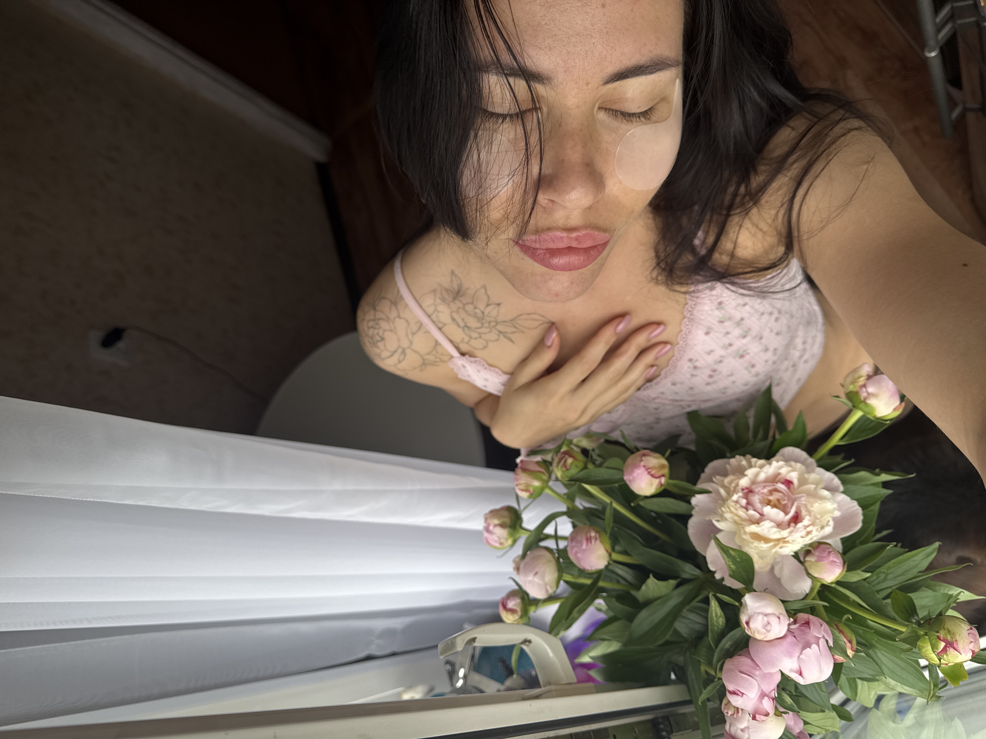 A young woman with dark hair and closed eyes is shown from a high angle, resting her head against a surface. She is wearing a light pink camisole with floral patterns and has a tattoo on her left shoulder. Under her eyes are clear patches. In front of her is a bouquet of pink peonies and green leaves. The background shows a white curtain to the left, and a wooden floor and textured wall to the right, suggesting an indoor setting. The image appears to be taken in Chaikovsky, Russia, as indicated by the context.