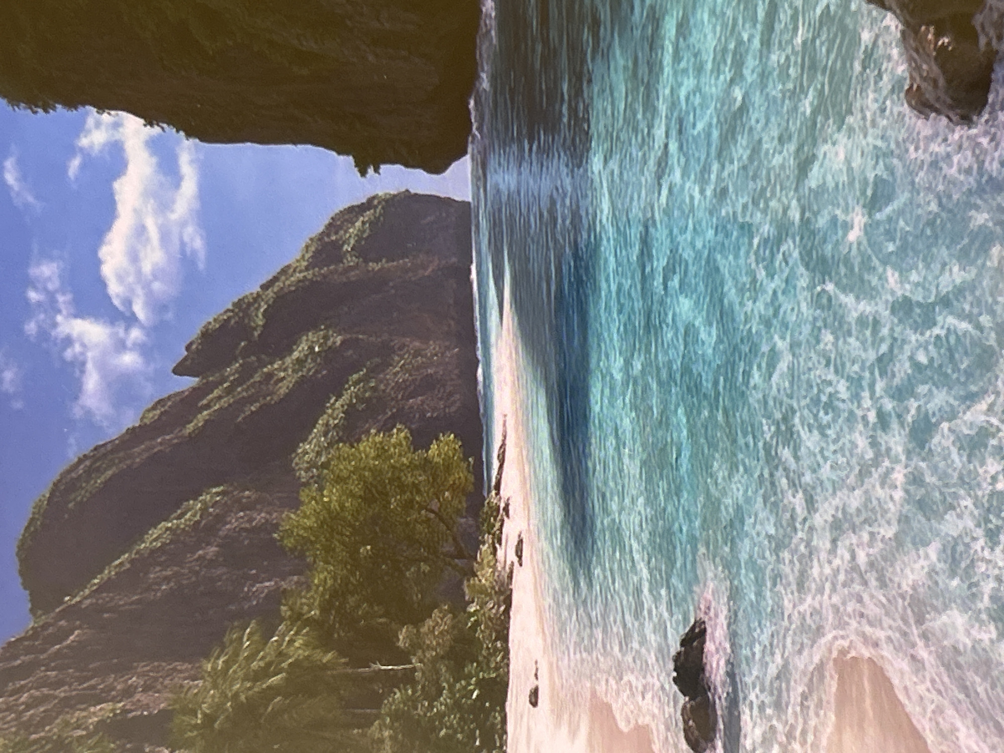A serene tropical coastal landscape is captured from a slightly elevated perspective. The dominant features are the exceptionally clear, turquoise and light blue ocean water, a pristine sandy beach, and dramatic, verdant mountains or cliffs.
The water, occupying the right and lower portion of the frame, exhibits different shades of blue and green, indicating varying depths and clarity. Gentle waves break onto the shore, creating white foam patterns. A light-colored, seemingly untouched sandy beach stretches along the base of the mountains, separating the water from the land.
On the left and in the background, towering mountains or steep cliffs rise majestically. These are densely covered in lush green tropical vegetation, including numerous trees and shrubs, suggesting a rich ecosystem. The texture of the foliage and rock formations is detailed. The sky above is a bright, clear blue with a scattering of fluffy white clouds. Some dark rocks are visible in the foreground water and at the very edge of the beach.
The overall impression is one of natural beauty and tranquility, indicative of a remote tropical paradise, likely an island or a secluded bay. The time of day appears to be bright daylight, with clear and sunny weather. There are no visible people, structures, or text in the image.