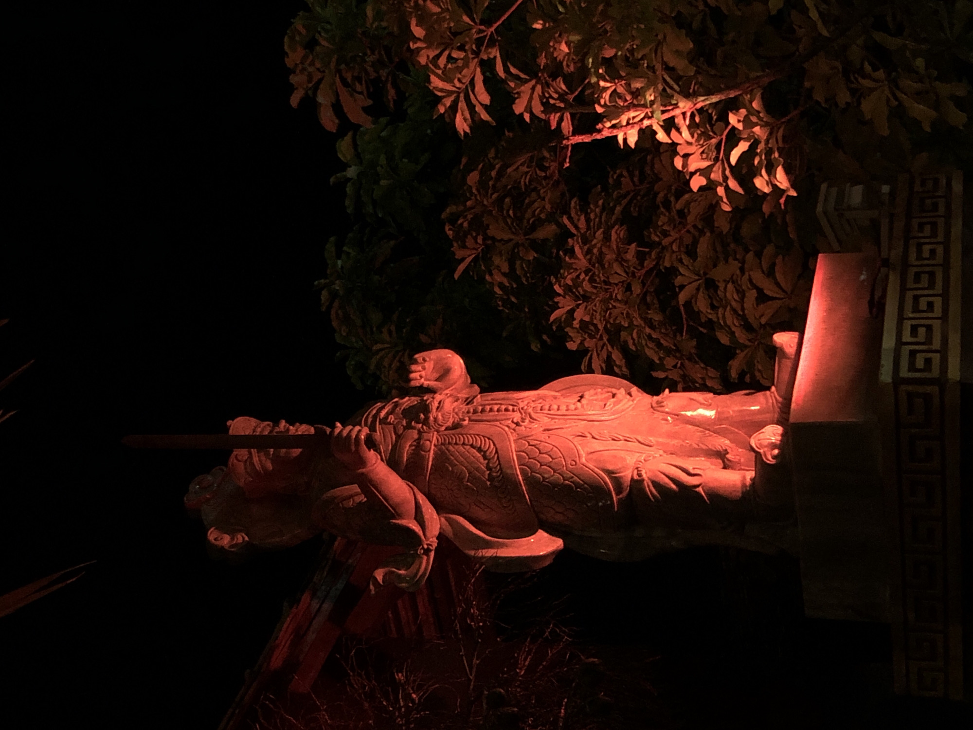 This is a low-angle, nighttime photograph of a large, red-hued statue of a warrior, possibly a deity or historical figure, in Mũi Né, Vietnam. The statue is illuminated by red light, giving it a warm, dramatic glow against the dark background. The warrior is depicted in a reclining or resting pose, with one arm resting on its body and the other holding a long, thin object, likely a sword or spear, horizontally in front of it. The statue is richly detailed, with intricate armor and facial features visible.
Behind the statue, a canopy of dense foliage is also lit by the red light, creating an eerie and atmospheric backdrop. The leaves are a mix of dark green and reddish-orange hues, suggesting the light source is artificial and casting an unusual color on the natural elements. To the right of the statue, a vertical column with a Greek key or meander pattern is visible, suggesting a decorative element of a temple, shrine, or other traditional structure. The overall scene is dark, with the main subjects sharply defined by the red lighting. The black sky and surrounding darkness indicate it is nighttime. There is no visible text in the image. The mood is solemn and perhaps awe-inspiring due to the scale of the statue and the dramatic lighting.