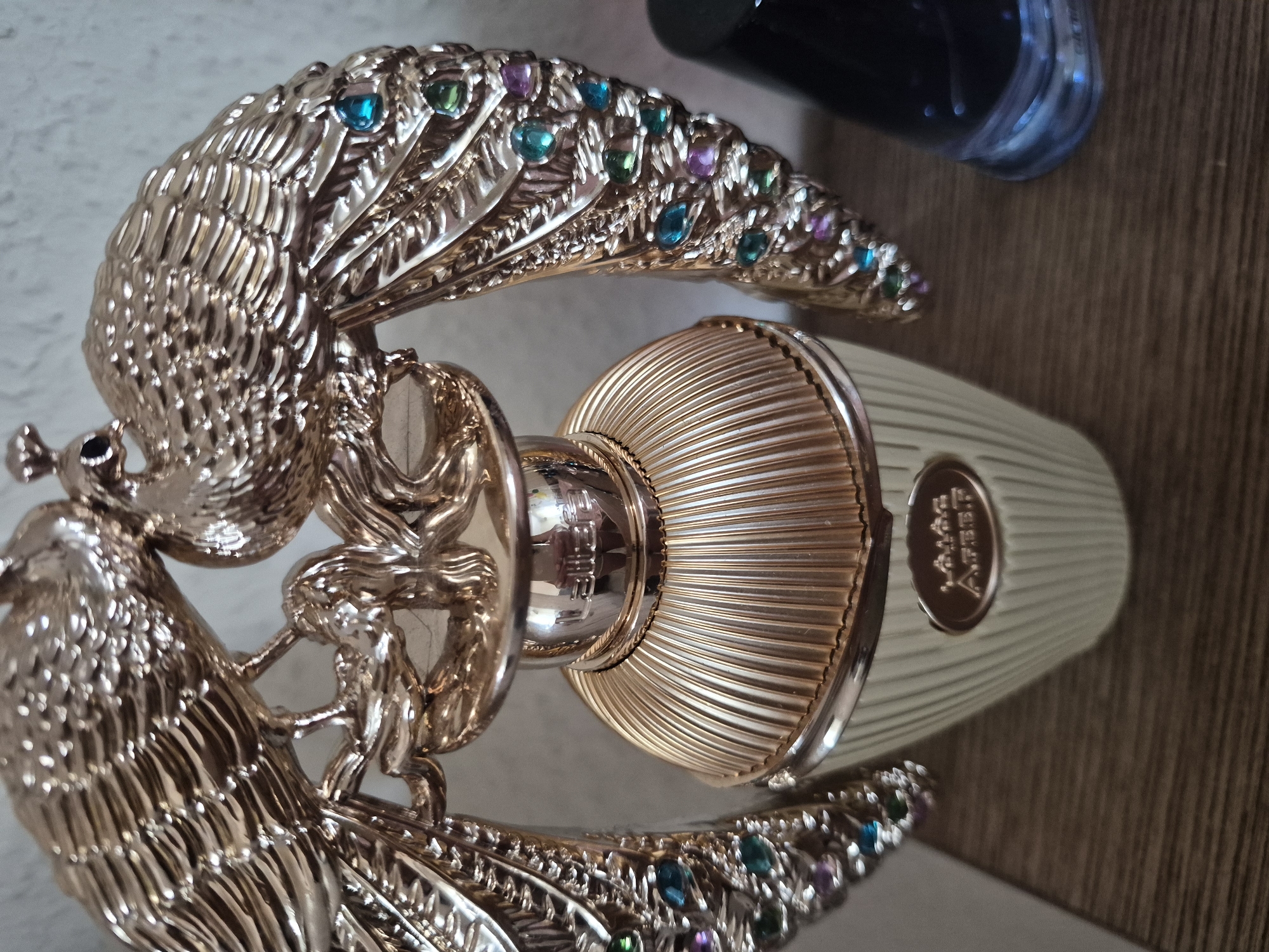 This is a close-up, eye-level shot of a decorative perfume bottle featuring two ornate, gold-colored peacocks perched on top of the bottle. The peacocks have detailed feathers and are adorned with colorful faux gemstones, including green, blue, and purple. The bottle itself is gold-toned with a ribbed texture and a round emblem on the front that appears to have Arabic script. The perfume bottle is positioned on a wooden surface, possibly a shelf or table. To the right of the perfume bottle, a dark-colored bottle, likely another cosmetic or perfume, is partially visible. The background is a plain, light-colored wall. The lighting is soft and diffused, creating subtle highlights on the metallic surfaces of the perfume bottle and peacocks.