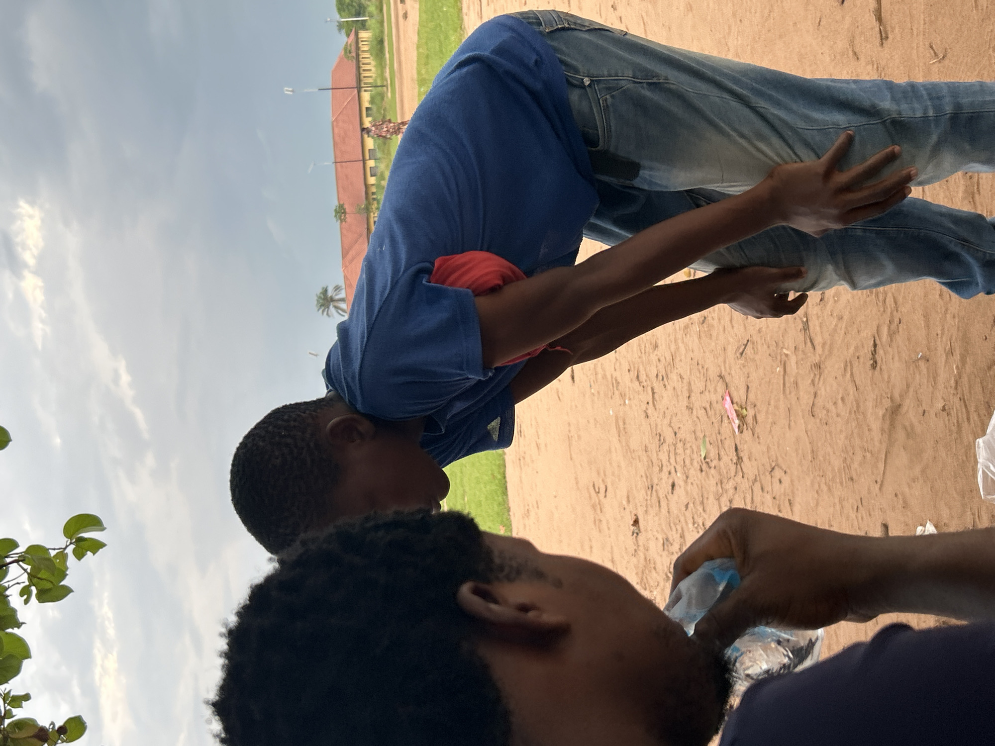 In Ihiagwa, Nigeria, two young men are captured in a moment under a cloudy sky. One man, in the foreground and facing away from the camera, is drinking from a plastic water bottle. He has dark, curly hair and is wearing a dark blue t-shirt. The other man, in the middle ground and facing left in profile, is bending over. He has short hair and is wearing a blue t-shirt over a red garment, and blue jeans. His hands are positioned on his hips and upper thighs. The ground is composed of sandy soil, scattered with a few pieces of debris. In the background, a long, low building with a terracotta-tiled roof and yellow walls is visible, along with a few palm trees and streetlights. The overall atmosphere is casual and informal, possibly in an outdoor recreational area or a semi-rural setting.