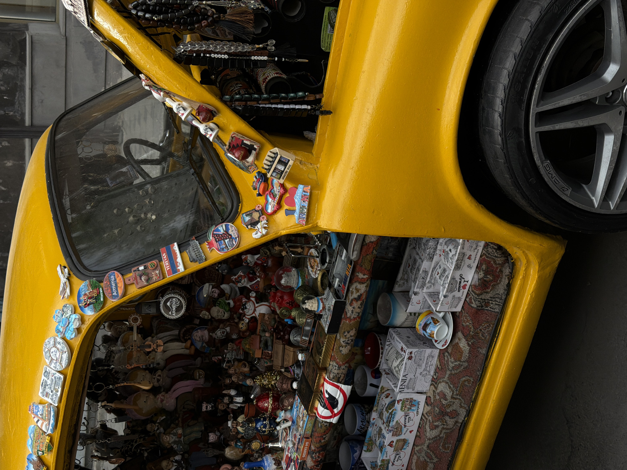 A bright yellow vehicle, appearing to be a custom-built display or art installation, is captured from a high-angle perspective, rotated 90 degrees counter-clockwise. No people are visible.
The scene showcases the vehicle, which is heavily adorned with various objects. The front section of the car (visible on the left side of the rotated image) has numerous souvenir magnets and pins affixed to its yellow body, particularly around the windshield. Through the windshield, the car's interior is densely packed with miniature figurines, dolls, and other small trinkets. The vehicle's hood (visible at the top-center) is open, revealing a compartment filled with beaded necklaces, bracelets, and other decorative items. A lower compartment or display area (visible on the right side) contains numerous white boxes, small cups, and more souvenirs arranged on a patterned rug. A large tire with an "AMG" logo on its sidewall is partially visible on the far right.
Notable details include the pervasive theme of Armenia: many magnets and boxes display "Armenia," "Yerevan," and "Echmiadzin," along with "I love Armenia" phrases. Other text includes "Armenian Wine" on a magnet and "54 CARDS" on a box. A symbol often associated with peace, a red circle with a line through a bird, is also visible. The tire shows "NOTIUS" and "225" in its markings. The overall impression is that of a unique souvenir stand or an art piece celebrating Armenian culture and tourist attractions.