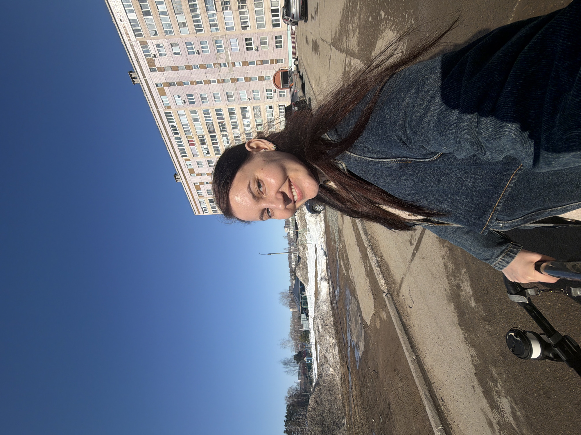 An outdoor scene in Chaikovsky, Russia, captures a smiling female individual in the foreground, facing the camera. She has long, dark brown hair and is dressed in a dark blue denim jacket over a light-colored garment. Her left hand is observed gripping the black handle of a wheeled object, possibly a stroller or bicycle, with a black and white cup holder attached. The middle ground features an asphalt and muddy dirt path with patches of melting snow. In the background, a large, multi-story residential building with a pinkish-beige facade and numerous windows dominates the right side of the frame, with several cars parked in front. To the far left background, a landscape of sparse trees and mounds of earth or snow is visible under a clear, bright blue sky. The overall environment suggests an urban area during early spring conditions.