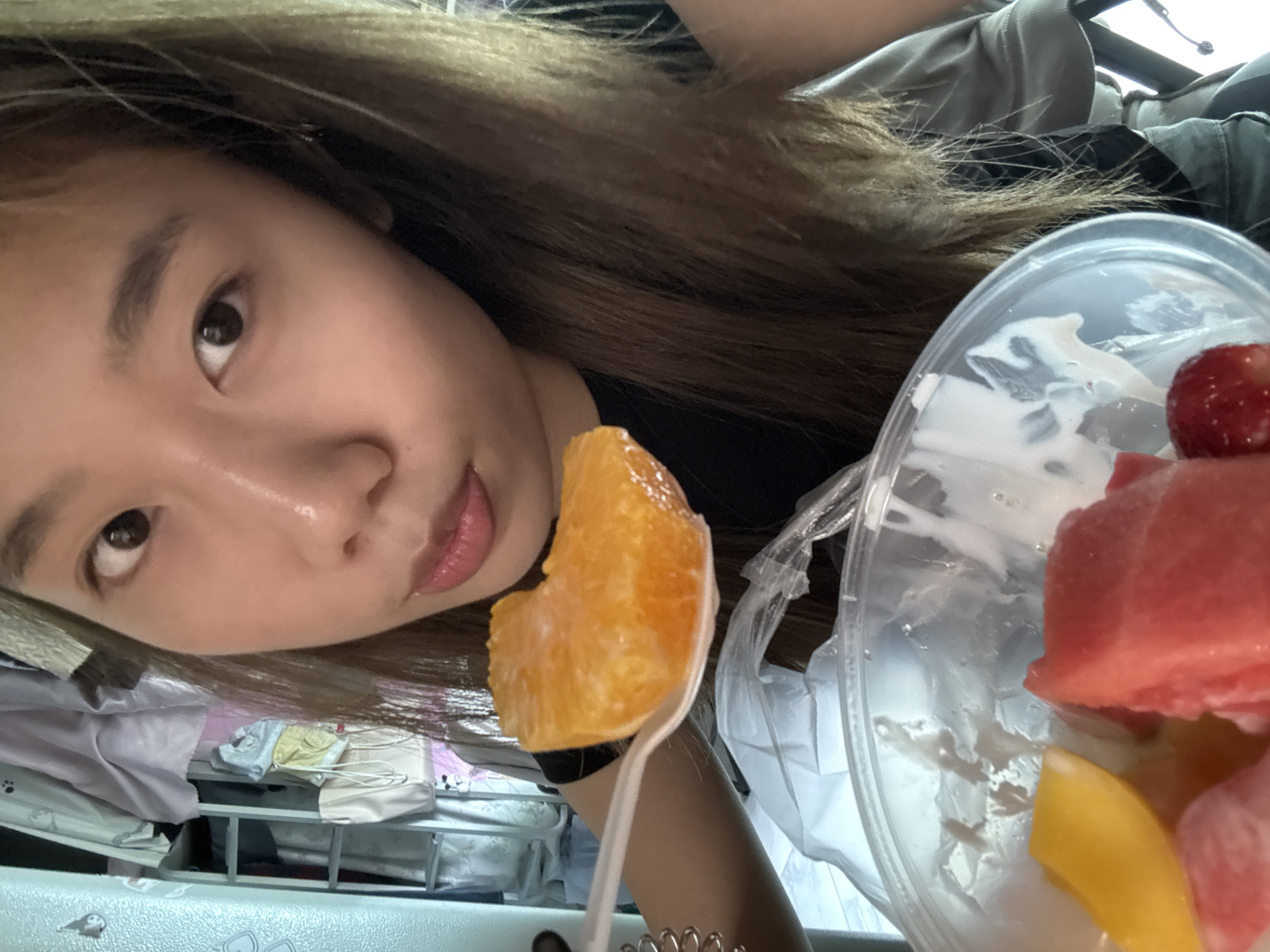 A person is holding a piece of orange on a plastic spoon towards the camera. A plastic container with watermelon, mango, and a strawberry is visible in the background. The person has brown hair and is wearing a black shirt. Bedding is visible in the lower left corner of the image. The location is Nan'an District, China.