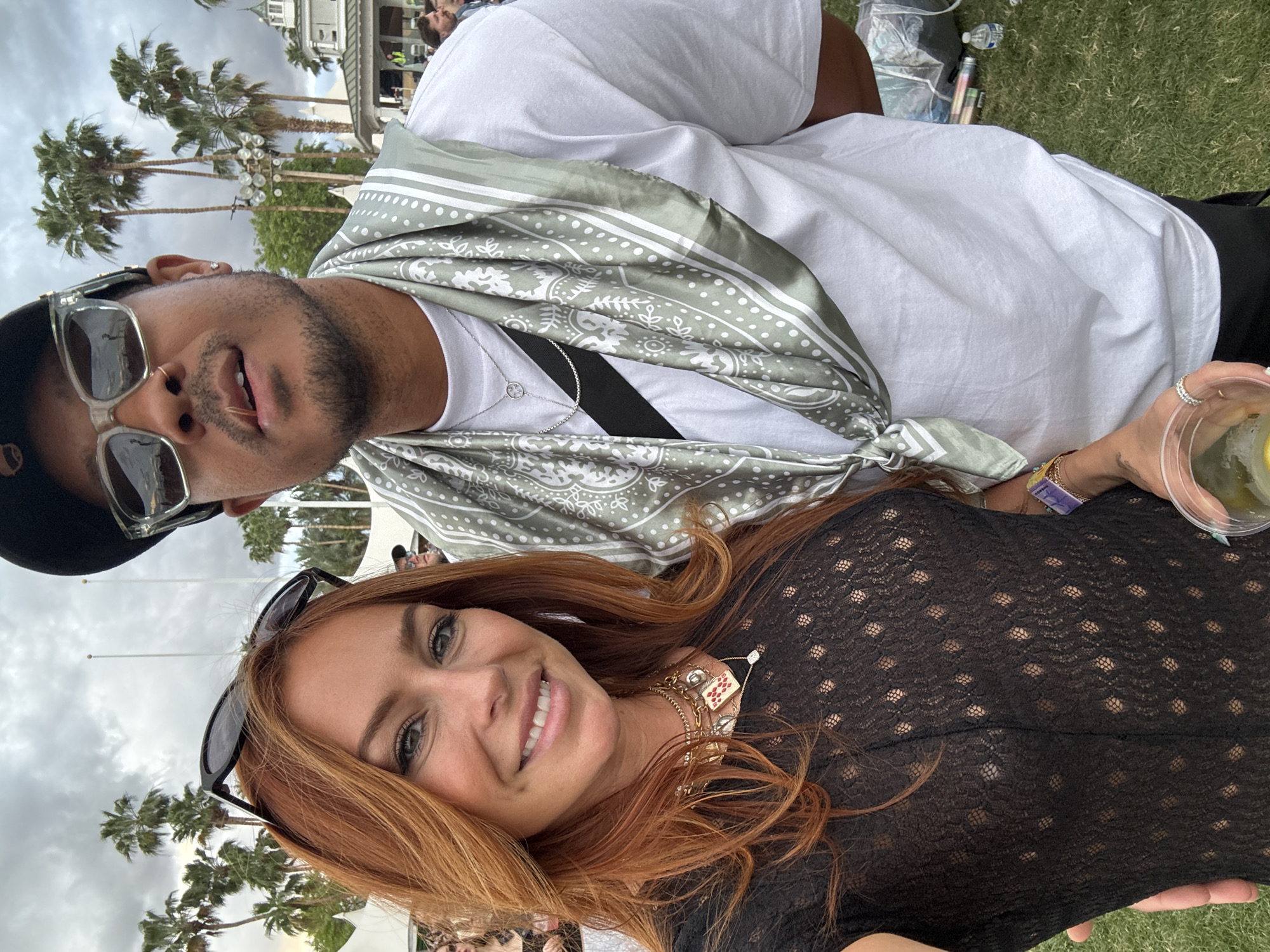 A selfie shows a woman and a man in La Quinta, California, United States. The woman is smiling with auburn hair, wearing a black shirt with a polka dot mesh pattern and necklaces and bracelets. She is holding a beverage with a lemon wedge. The man is above her, wearing a black baseball hat, sunglasses, a white t-shirt, and a bandana tied around his neck. Palm trees and a building are visible in the background.