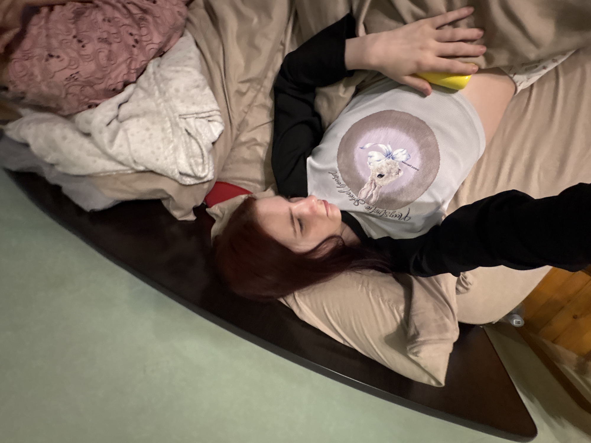 The image shows a young person lying in a bed, viewed from a high angle. The person has reddish-brown hair and is wearing a light-colored t-shirt with a graphic of a bunny and text that appears to read "Moonstone School Board". The t-shirt has black sleeves. The bed is covered with tan-colored sheets and a patterned pink blanket. The background shows a wall painted a light green color. The image appears to have been taken in a residential setting, likely a bedroom. There is no indication of the specific location in Sinyavino, Russia.