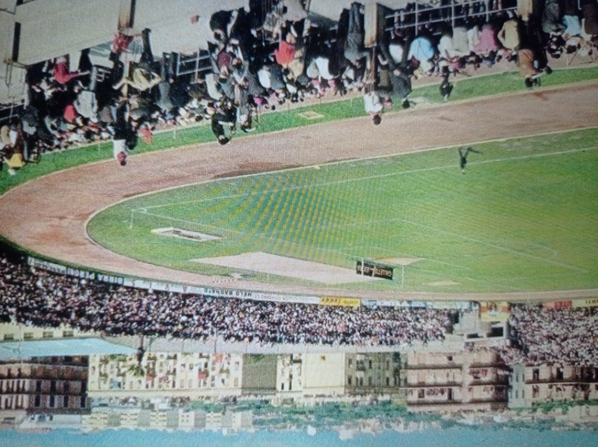 This is a wide-angle, slightly tilted, color photograph of a crowded outdoor stadium, likely a soccer match, with a cityscape visible in the background. The stadium features a green grass field with white markings, and a dirt track surrounding it. Dense crowds of spectators fill the tiered seating on both sides of the frame. Many individuals in the foreground and midground are wearing casual clothing, and some are standing or leaning on railings. A lone figure is walking on the track in the midground. In the distance, buildings of varying heights, some residential and some potentially commercial, form a sprawling urban landscape under a hazy sky. The image's quality suggests it may be an older photograph due to its slightly grainy texture and muted color palette. The location is not identifiable as Mukachevo, Ukraine, based on visual cues within the image.