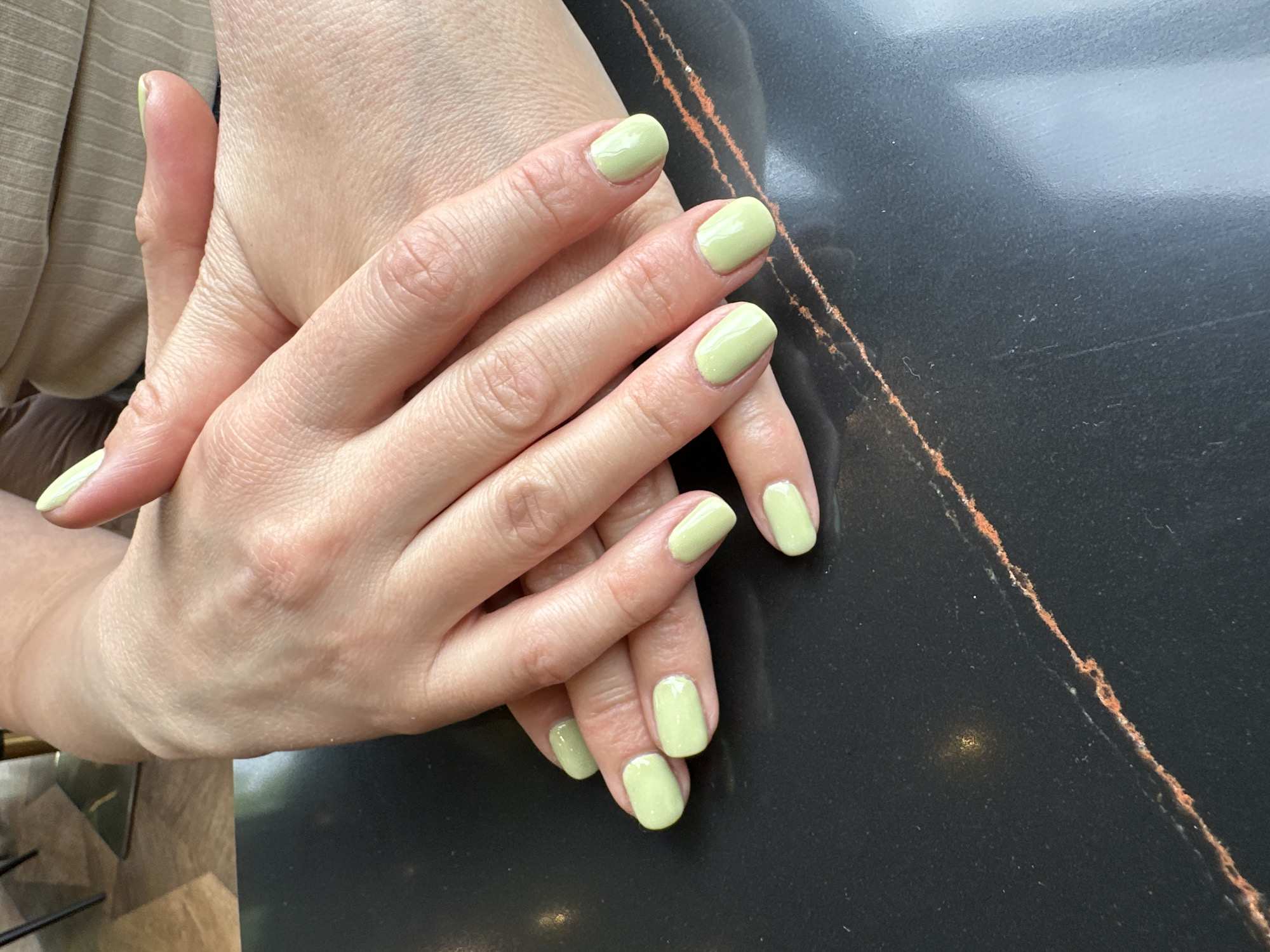 The image shows a close-up of two hands with manicured fingernails painted in a light green color. The hands are resting on a dark, possibly black marble surface with a copper-colored vein running through it. The hands appear relaxed and are intertwined. Part of the person's light brown shirt is visible in the upper left corner. The setting appears to be indoors. There is no indication of Montana, Bulgaria, or any specific location within that city.
