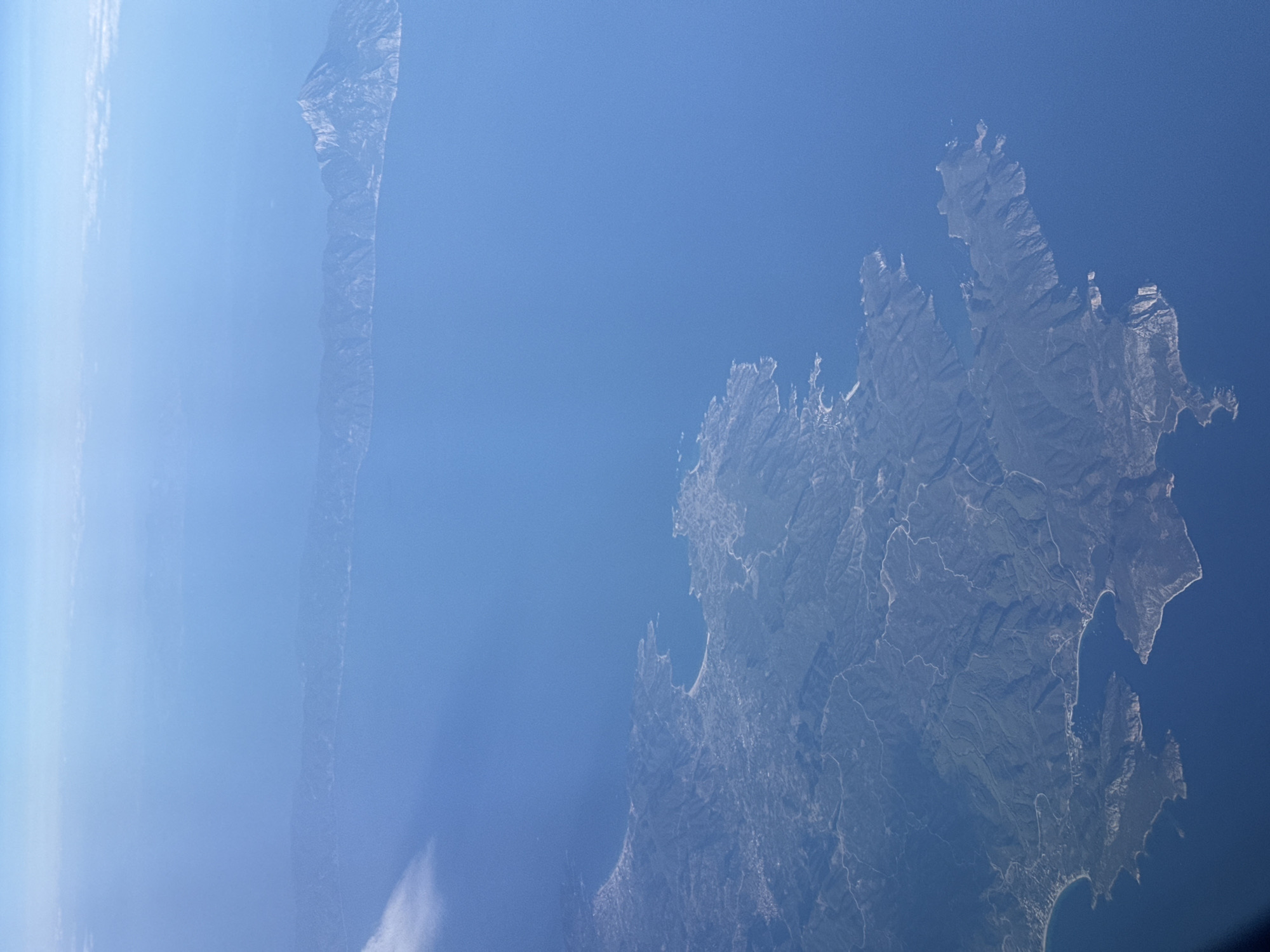 The image is an aerial view of a coastline with mountains and the sea. The water is a deep blue, and the land is covered in green vegetation with some visible roads and settlements. In the distance, a larger landmass with a prominent mountain peak is visible across the water. The sky above is clear and blue, with some wispy clouds. The image does not depict the city of Lukovit, Bulgaria, as Lukovit is an inland city and not located on a coastline.
