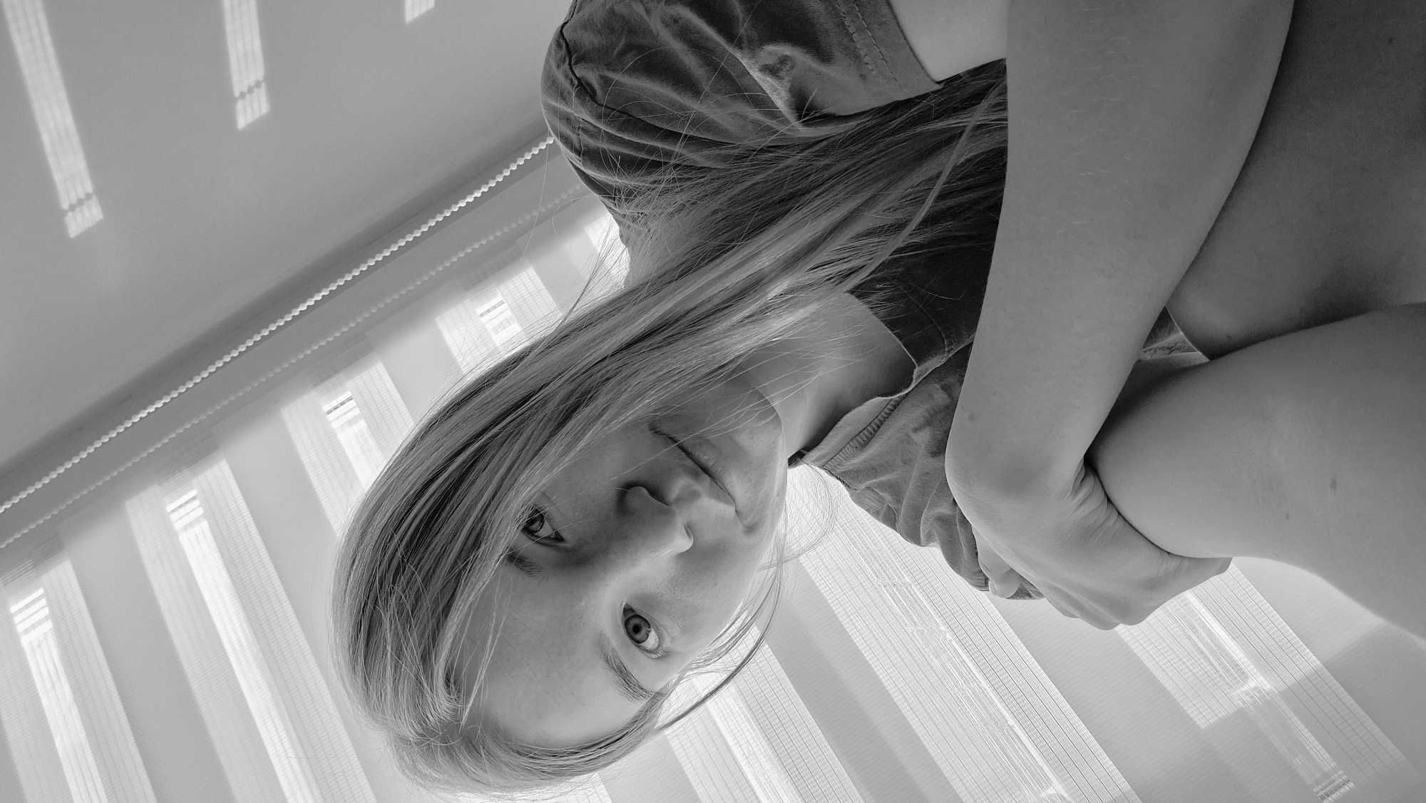 A monochrome, high-angle shot depicts a young woman with light hair, likely in an indoor setting. She is looking up at the camera. She has her arms crossed and is wearing a t-shirt. Blinds are visible in the background. The location is not discernible from the image.
